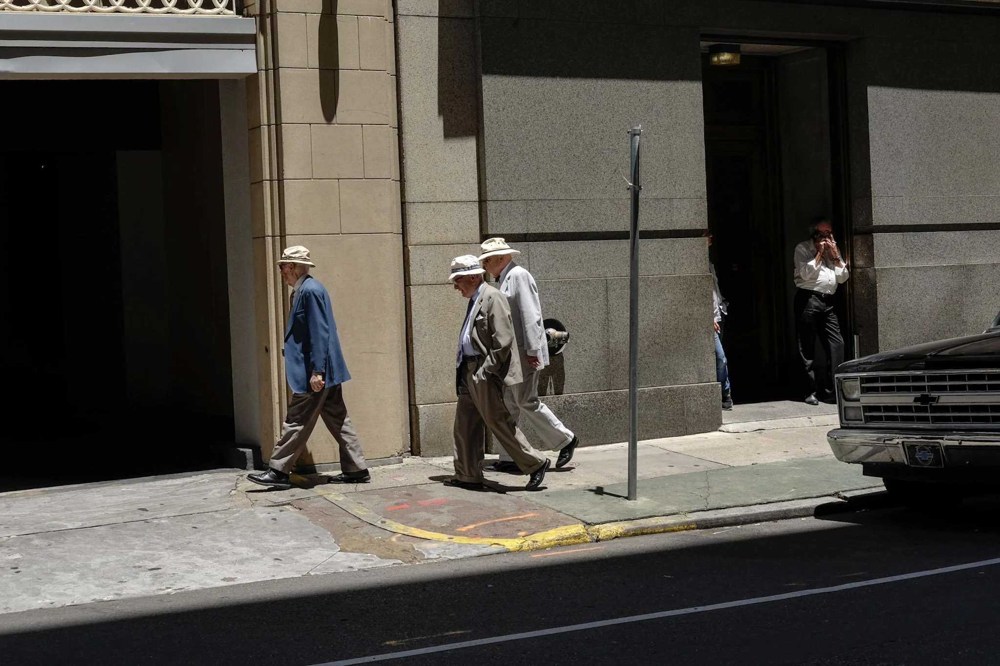 Four elderly men walking on a sidewalk in front of a building with beige stone walls, wearing hats and light-colored suits. A person is taking a photo through a doorway, and a car is partially visible on the right side.