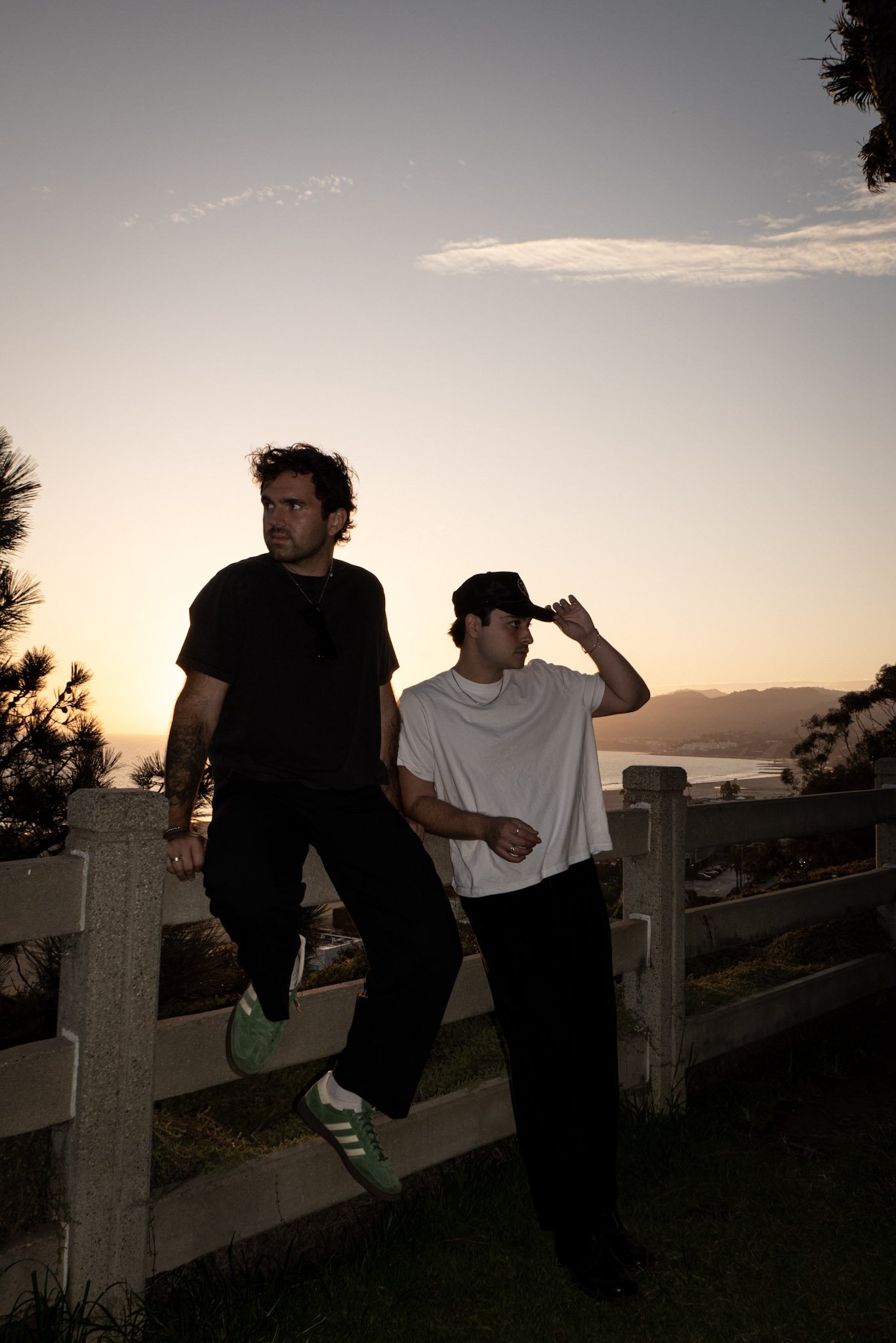 Two young men posing outdoors during sunset, with one sitting on a fence and the other standing. The background includes a scenic view of water and distant mountains.