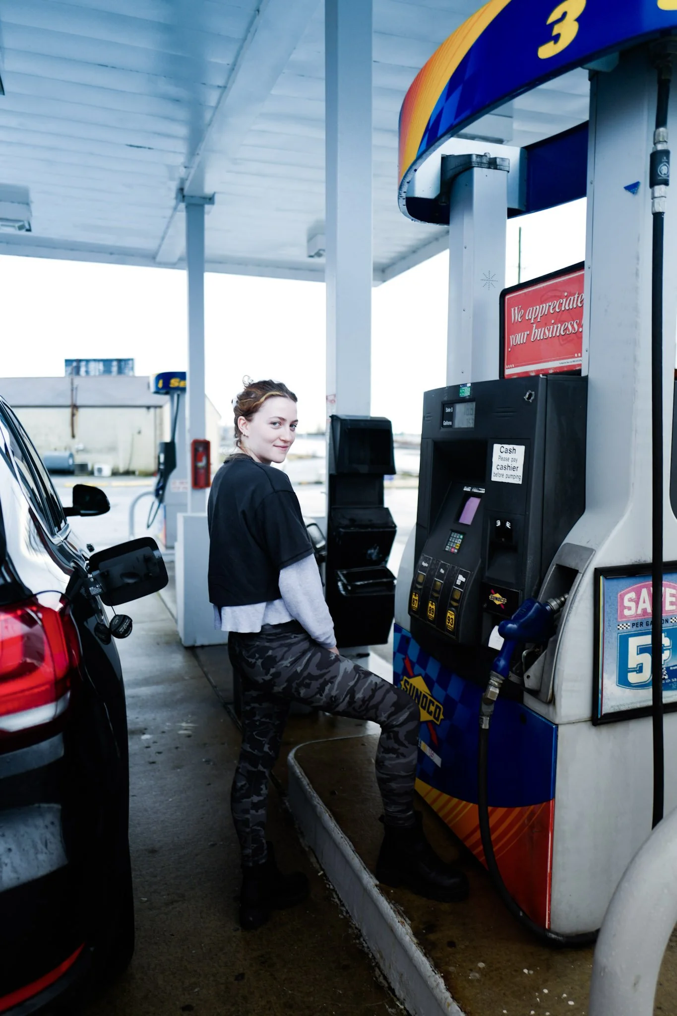 A young woman with brown hair, wearing camouflage pants, a black jacket, and black boots, is sitting and filling her car with gas at a gas station. She is looking at the camera and smiling. A red vehicle is parked nearby.