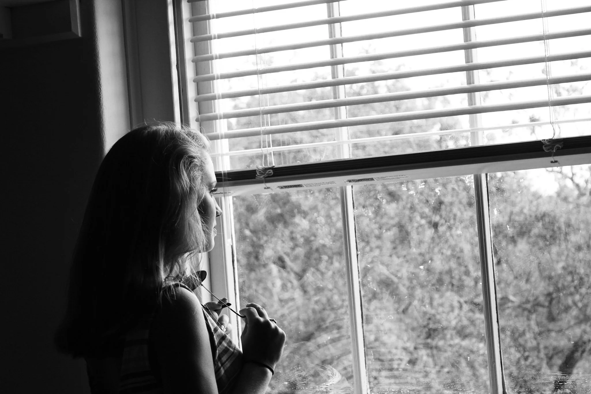 A woman with long hair looking out a window with blinds, using her phone, in black and white.