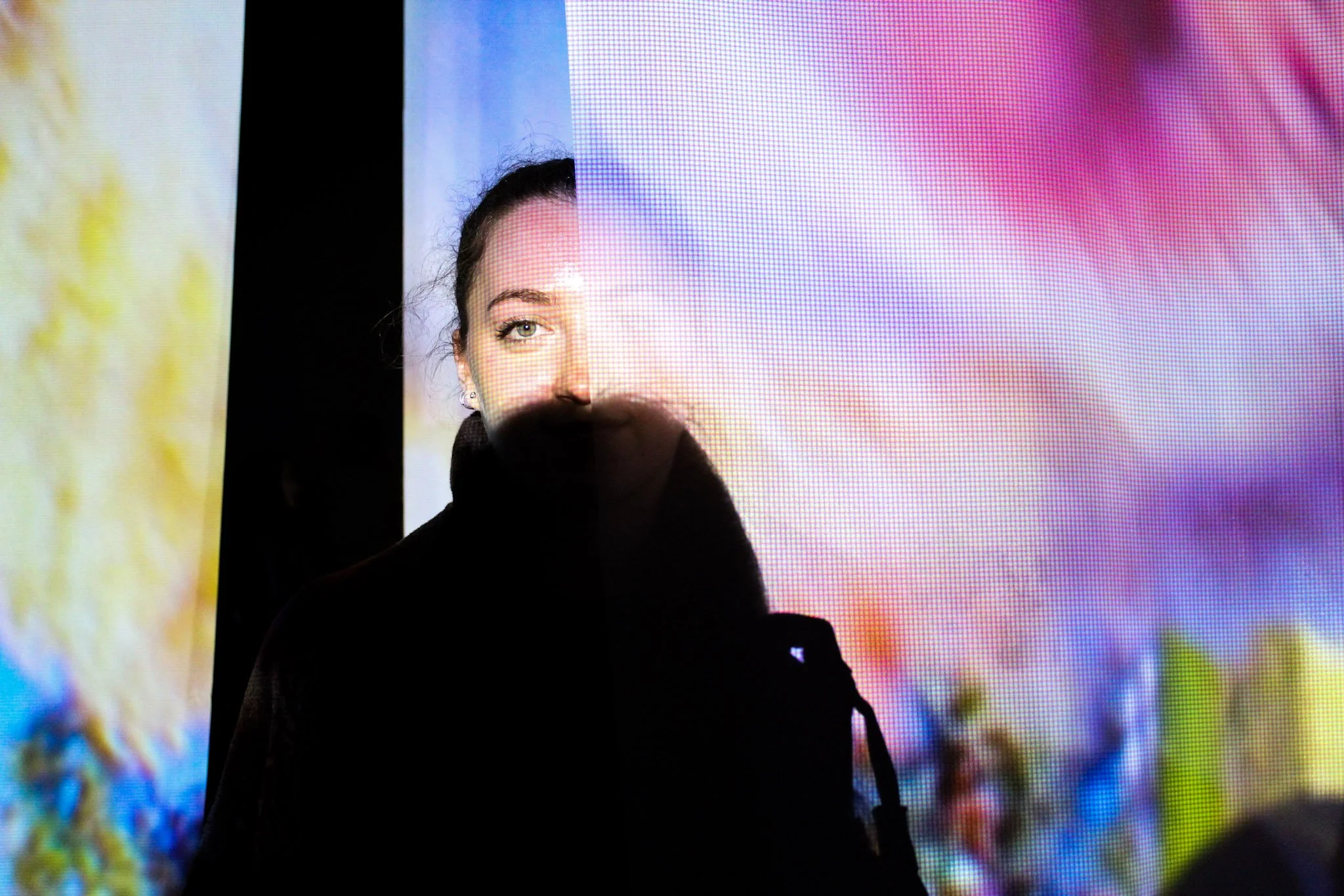A person with dark hair and earrings standing in front of a colorful, pixelated digital screen, partially obscured by a black shape.