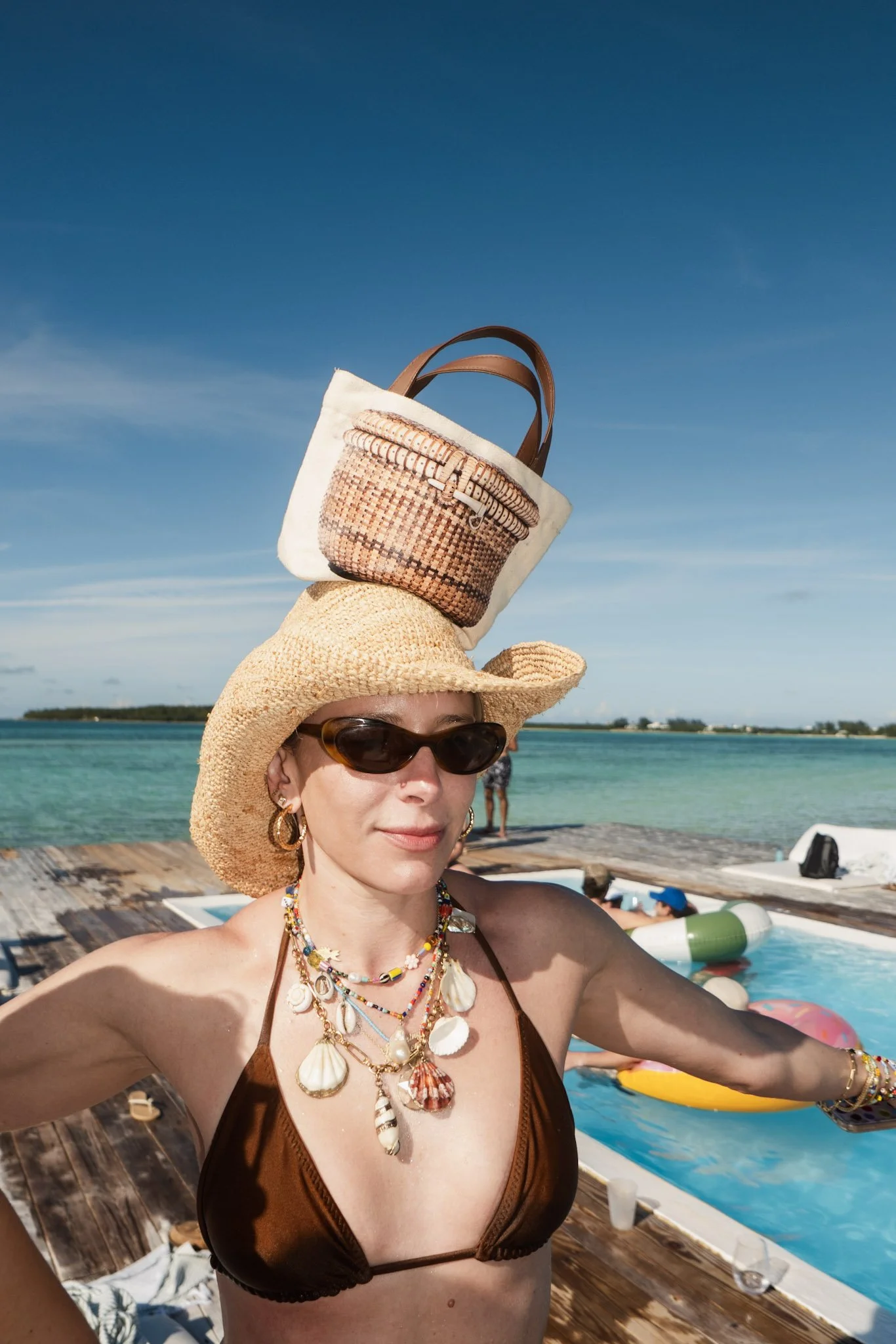 Woman wearing sunglasses, a straw sunhat, and multiple shells and beaded necklaces, taking a selfie at the beach with the ocean and clear sky in the background.