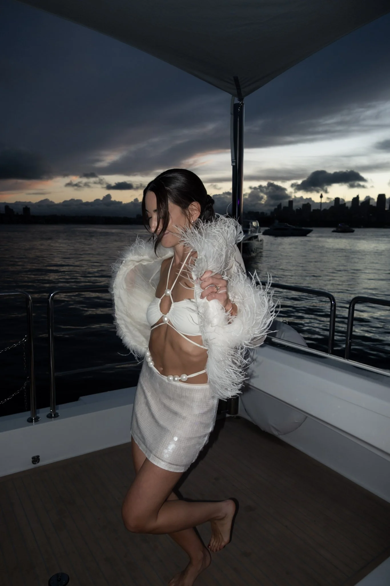 A woman dressed in white on a boat, smiling with her eyes closed, during sunset with a city skyline and water in the background.