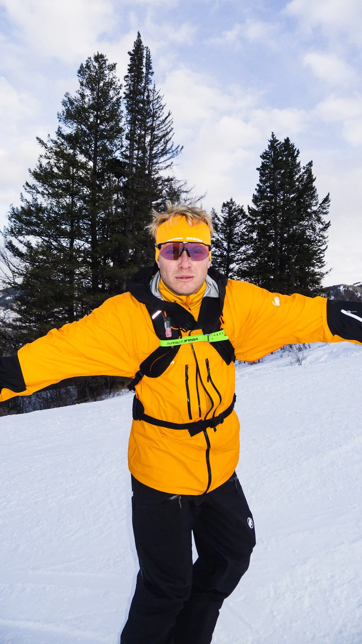 A man wearing a bright yellow jacket, black pants, and a yellow headband stands with arms outstretched in a snowy landscape with pine trees and a cloudy sky behind him.