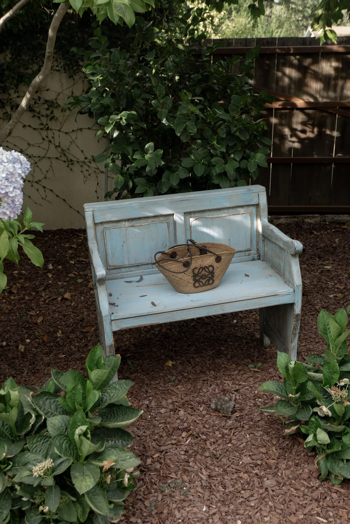 A weathered light blue wooden garden bench with a small woven basket on it, set in a lush garden area with green plants, flowers, and a dark wooden fence in the background.