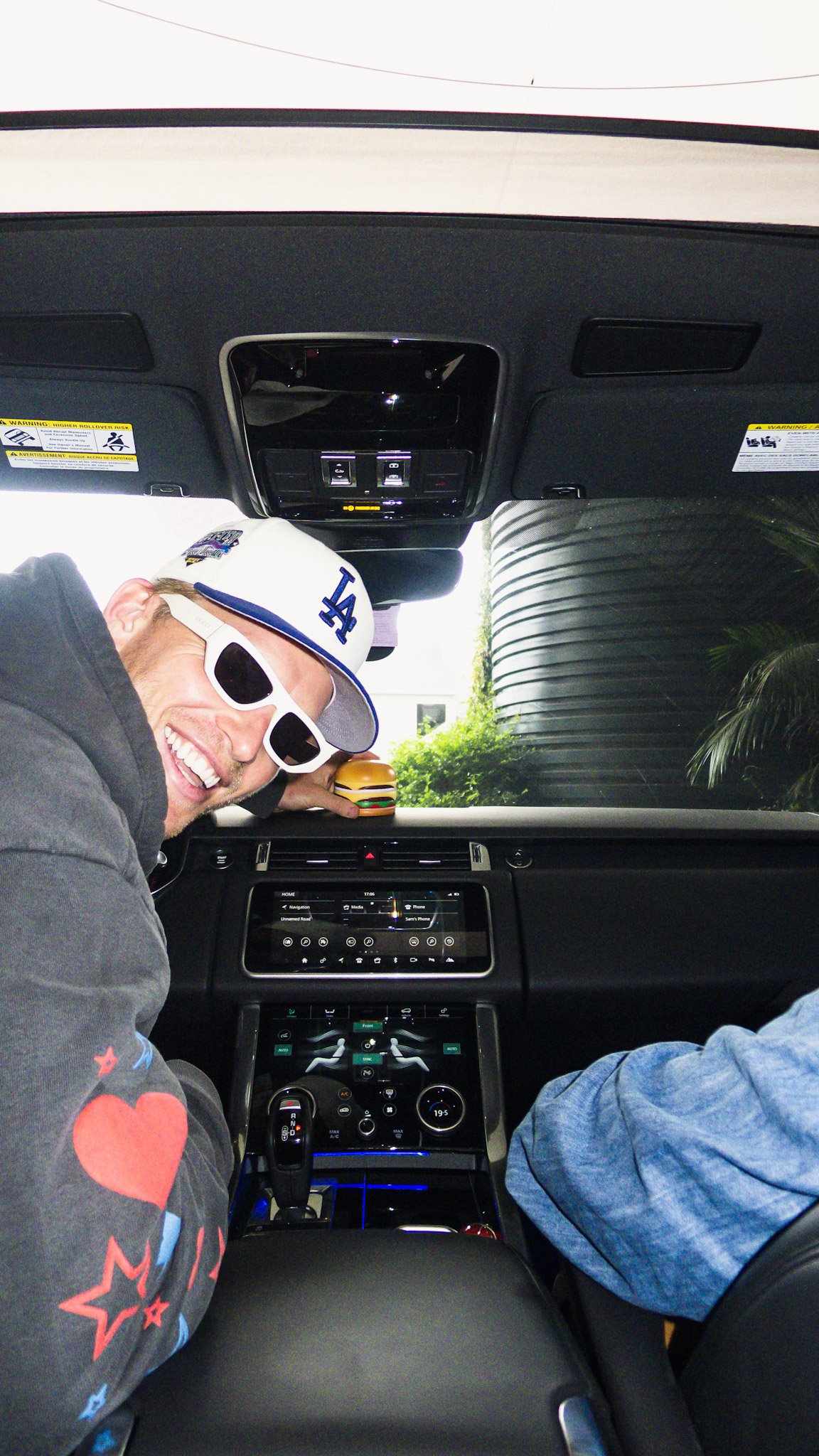 Inside a car, a man with sunglasses and a Los Angeles Dodgers cap is smiling, sitting in the front passenger seat, with a small hamburger toy on the dashboard and another person's arm partially visible on the right.