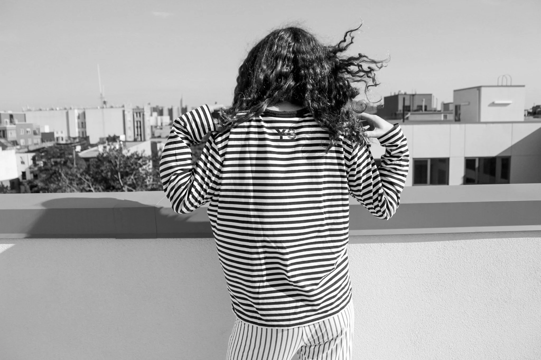 A person with curly hair wearing a striped Y-3 shirt and striped pants standing on a rooftop overlooking a cityscape in black and white.