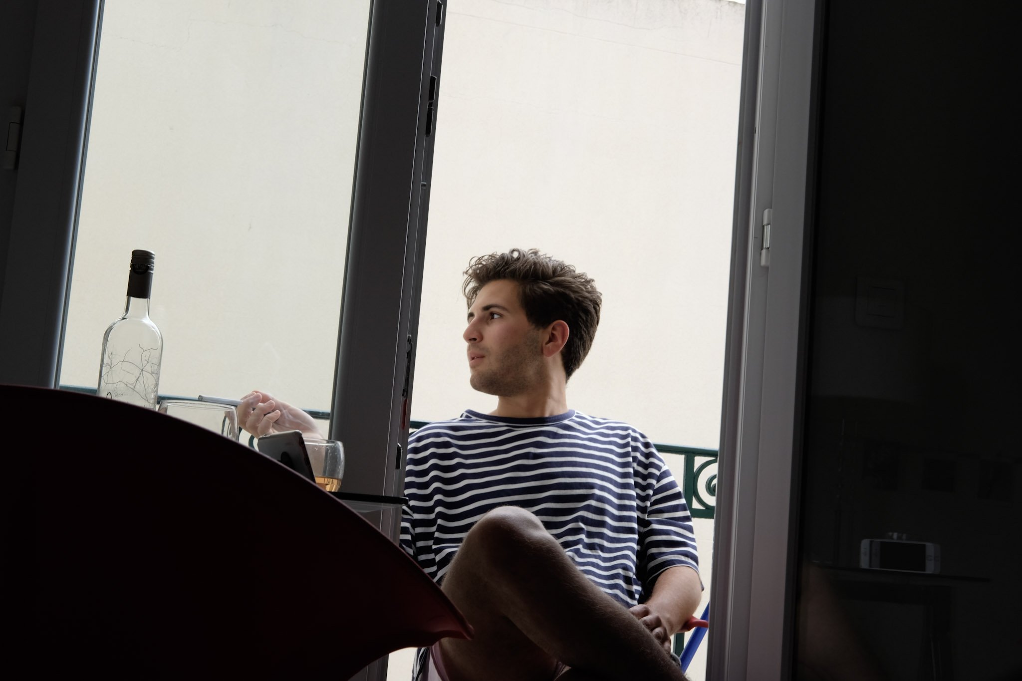 A young man with dark hair in a striped shirt sitting at an outdoor table, looking to his left, with a bottle of alcohol, a glass, and a smartphone on the table.