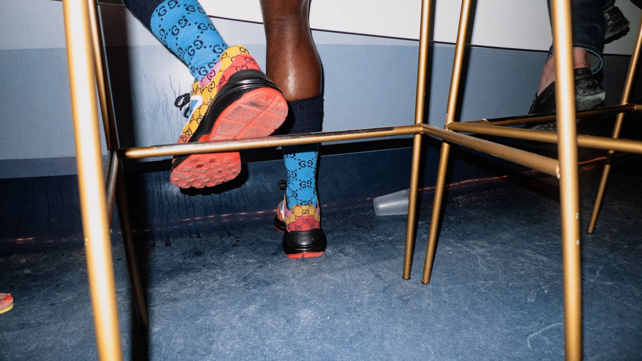 View of a person standing under a table, showing their legs and colorful Gucci sneakers with a bright red sole. The person is wearing a blue sock with black text and a yellow sock with black Gucci logos. The setting appears to be indoors with a blue 