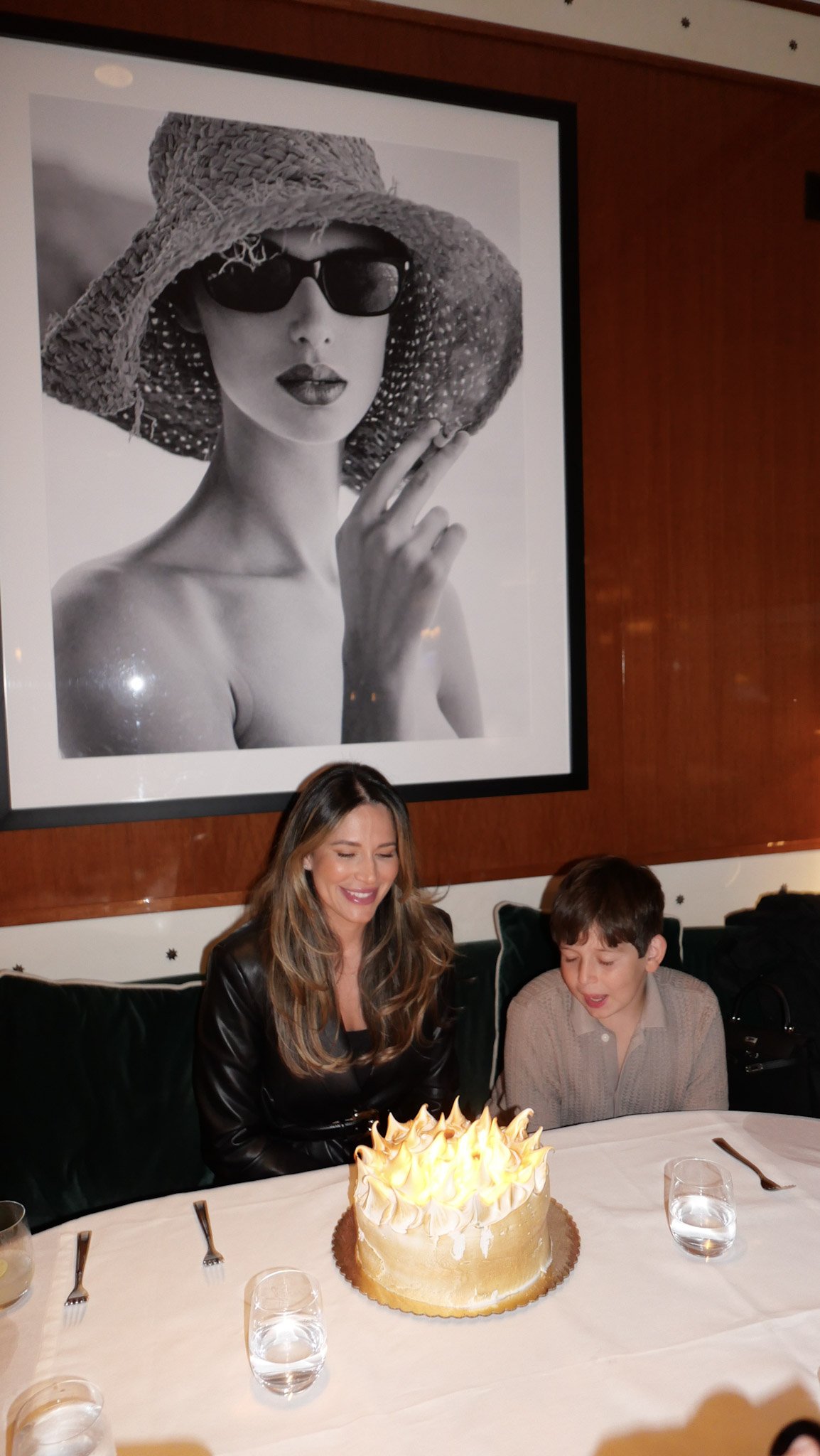 A woman and a boy sitting at a table with a birthday cake with flames, in a restaurant. There is a large black-and-white portrait of a woman wearing sunglasses, a wide-brimmed straw hat, and lipstick on the wall behind them.