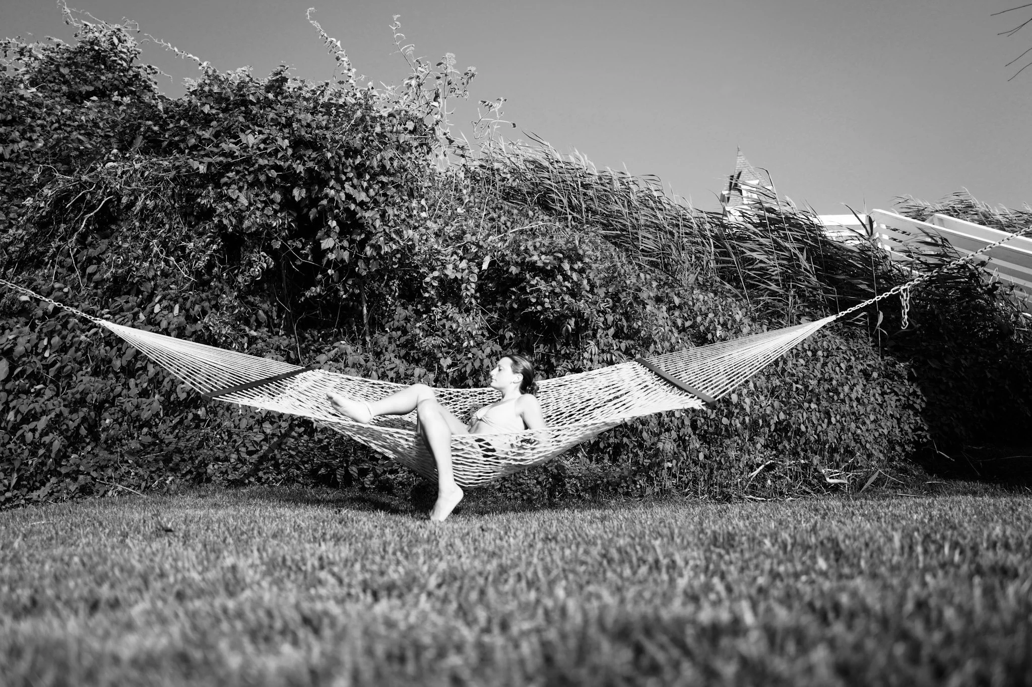 A woman relaxing on a hammock in a backyard surrounded by dense bushes and tall grass, under a clear sky.