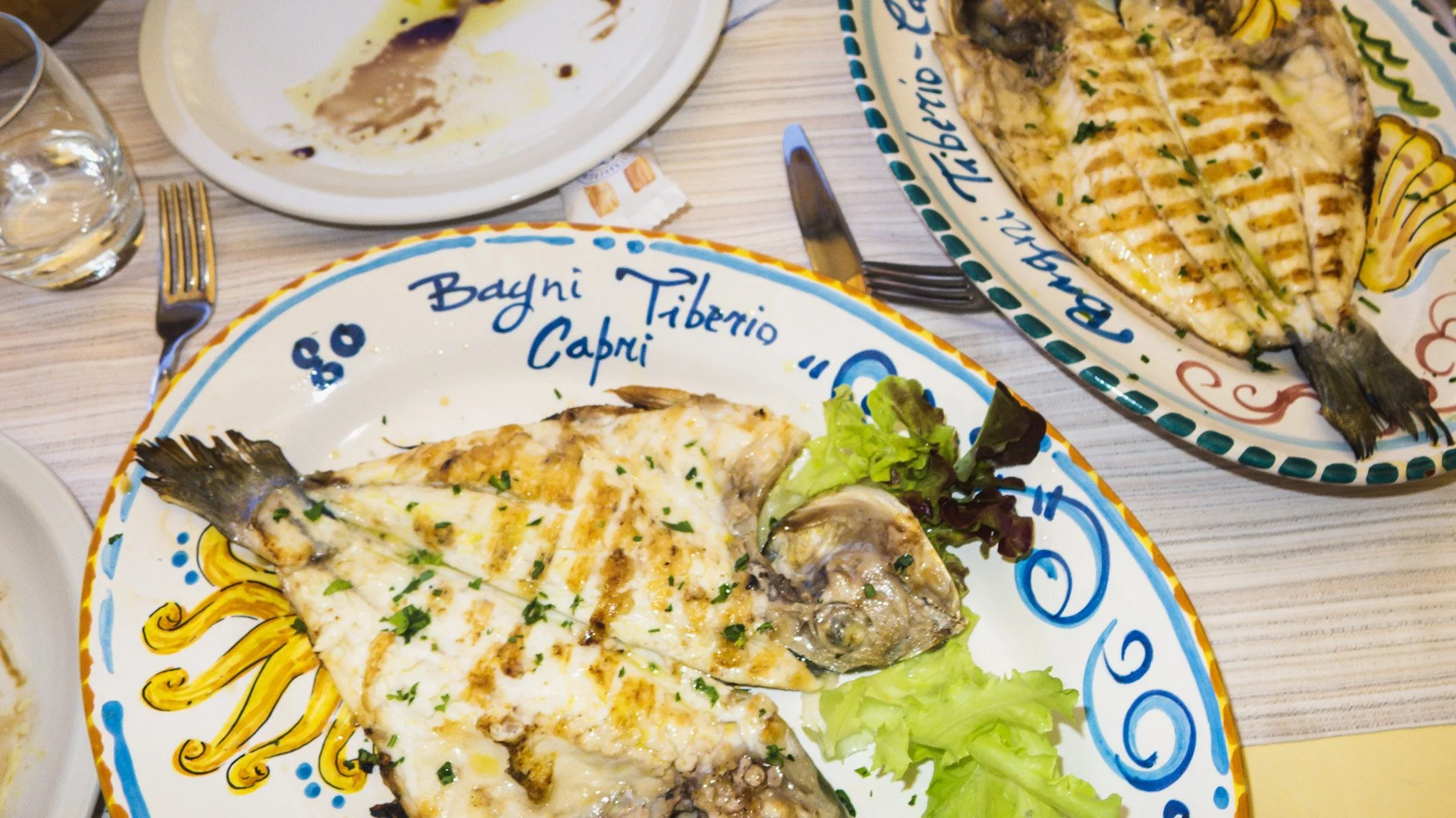 Plate with grilled fish, garnished with chopped herbs, with lettuce on the side. Another plate with more grilled fish and some grilled vegetables is in the background, on a colorful decorative plate.