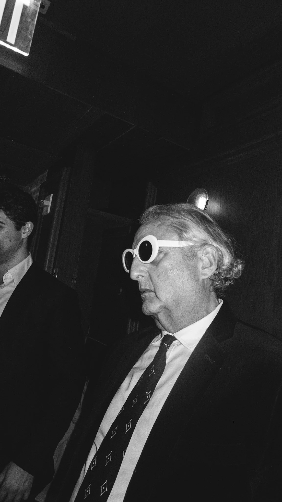 An older man wearing sunglasses and a suit, sitting next to another person, in a dimly lit interior setting.