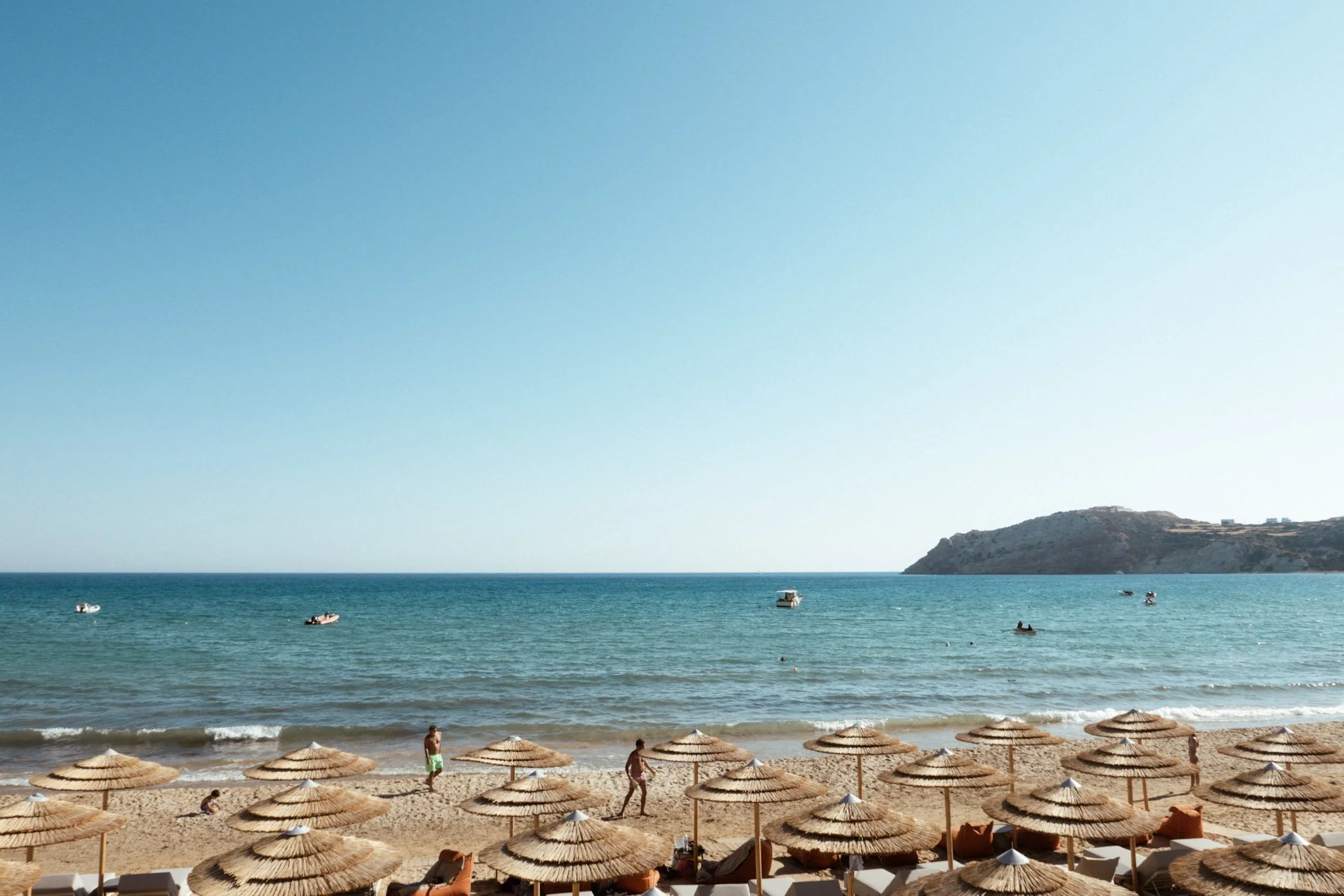 Beach scene with straw umbrellas, people walking, and calm water with boats and a hillside in the background.