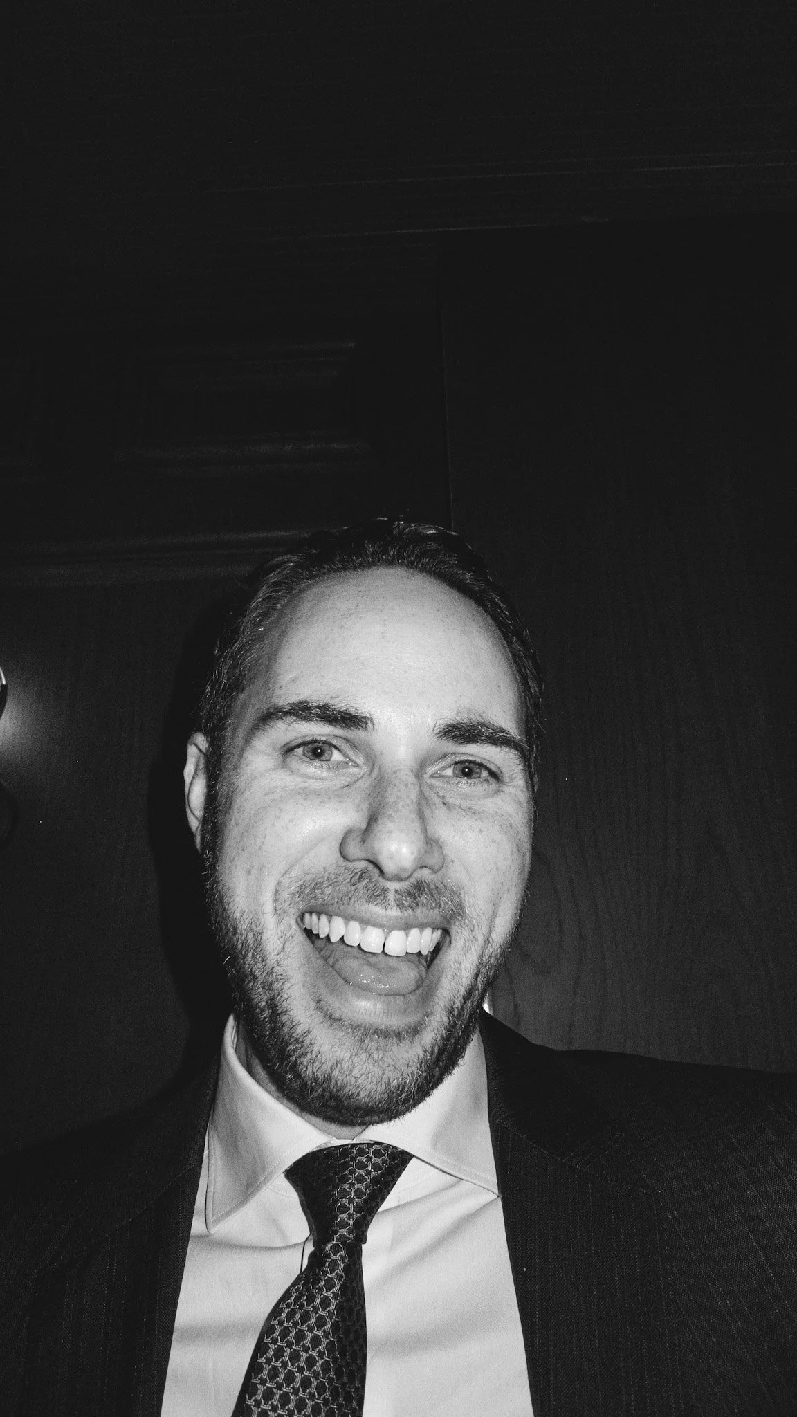 A black and white photo of a smiling man wearing a suit and tie, inside a room with a dark ceiling and a wall in the background.