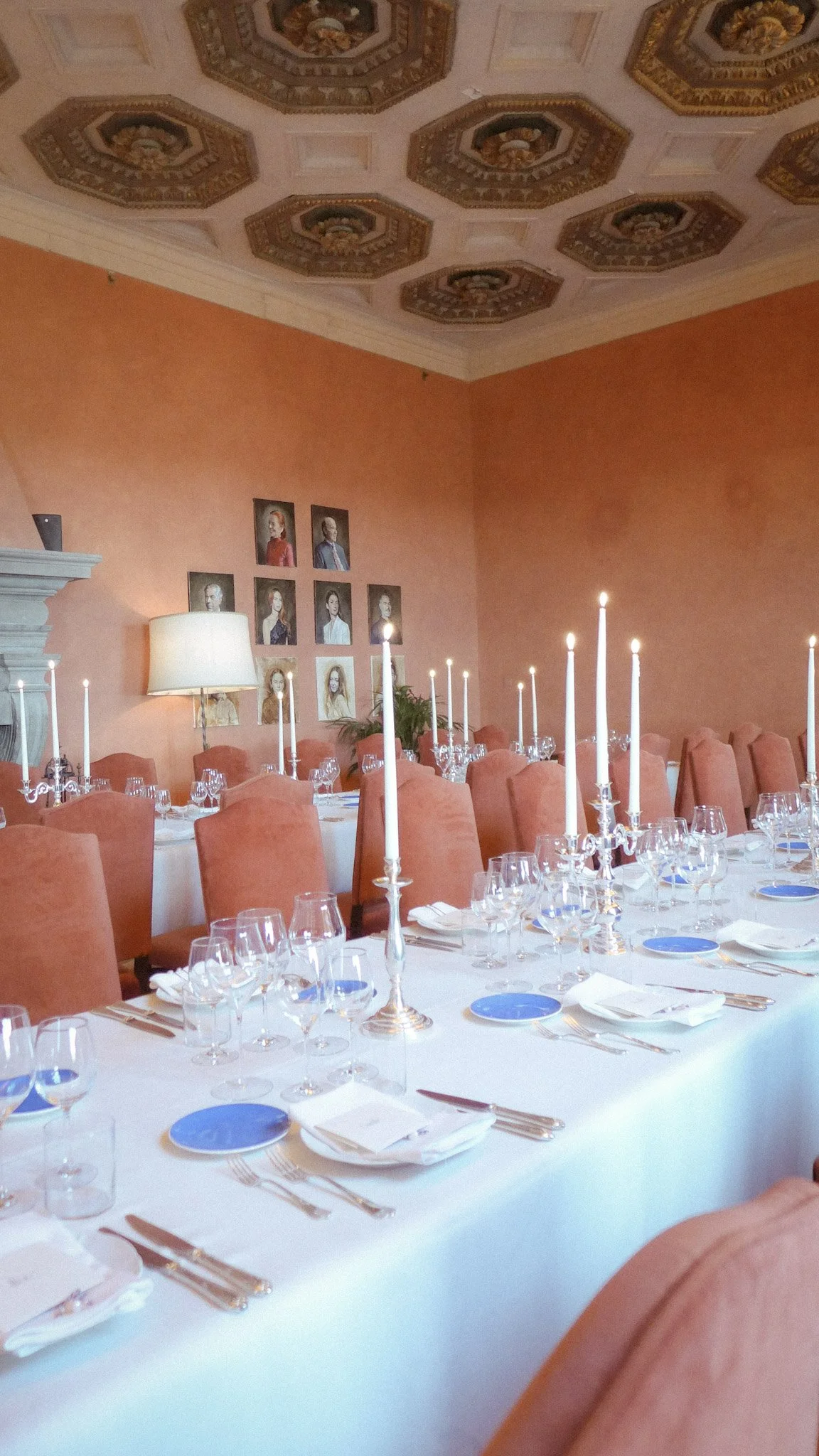 Elegant dining room with a long table set with glassware, silverware, plates, candles, plush pink chairs, paintings on the peach-colored wall, a lamp, and a decorated ceiling.