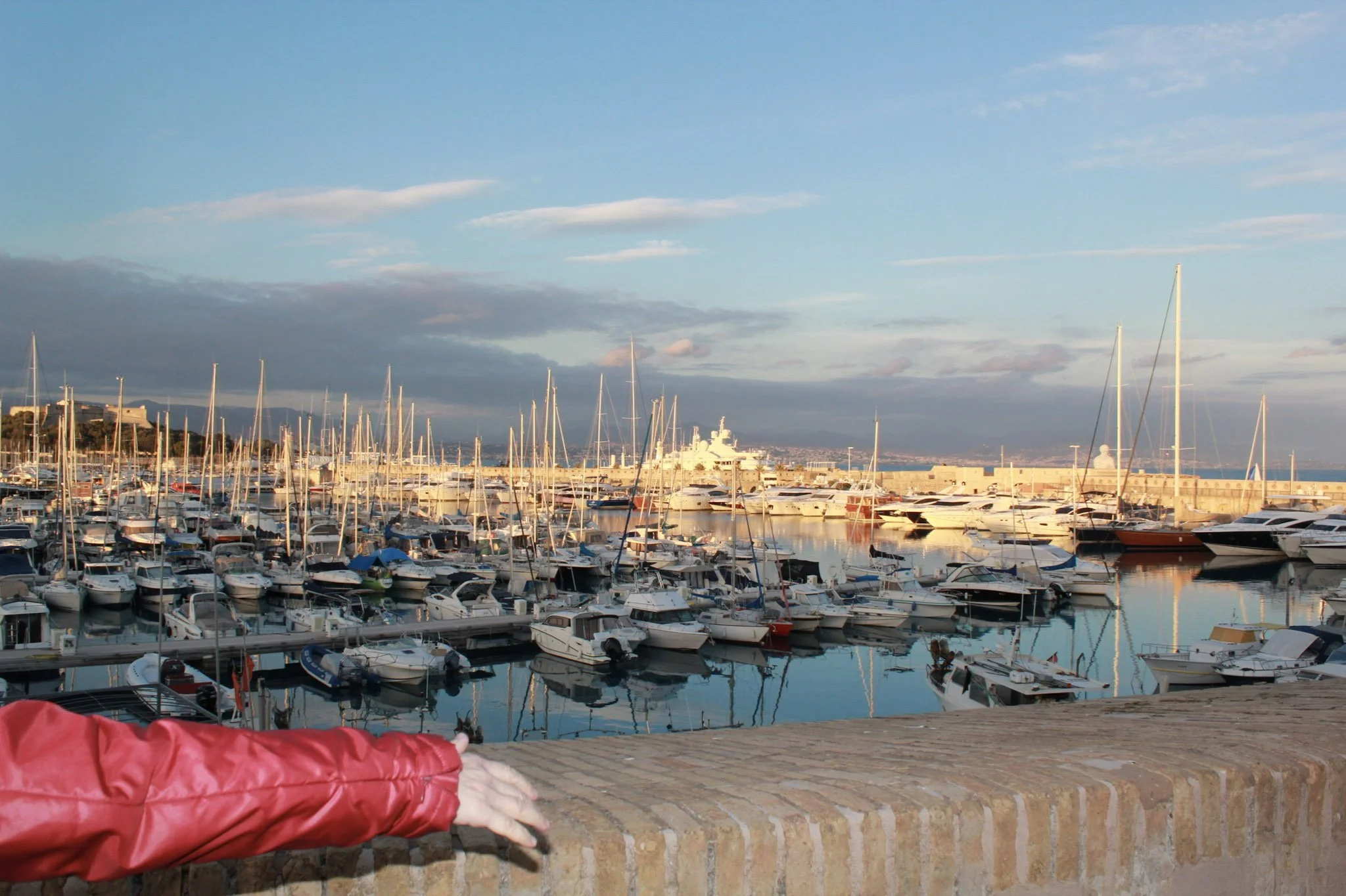 A person's arm in a red jacket resting on a stone wall overlooking a marina filled with yachts and sailboats, with mountains and clouds in the background.