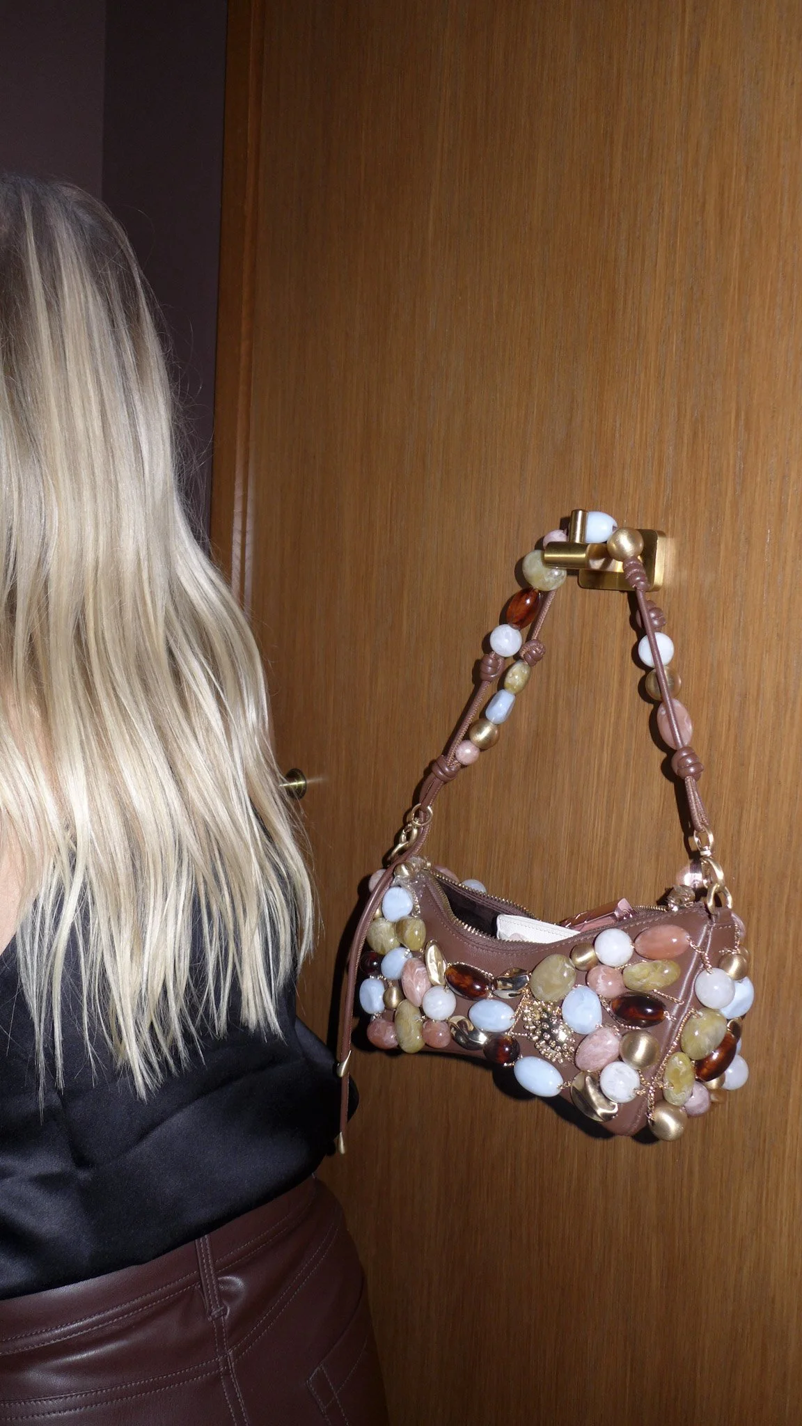 A woman with long, wavy blonde hair standing in front of a wooden door with a beaded handbag hanging from a hook on the door.