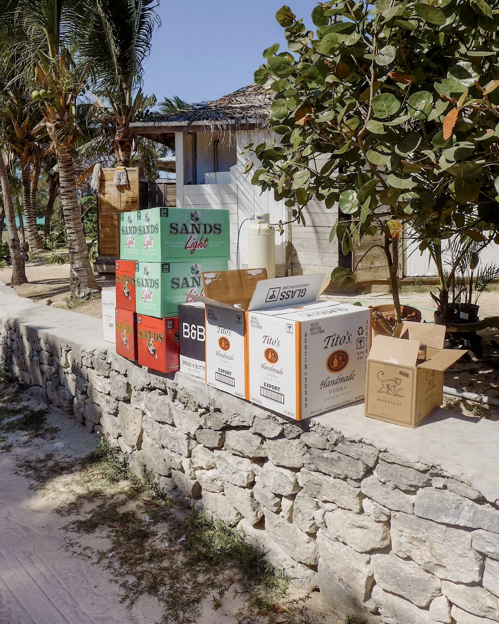 Beach scene with a stone wall, palm trees, and a small white hut with a thatched roof, boxes of drinks, and a sign reading 'Glass' and 'Handmade' in front.