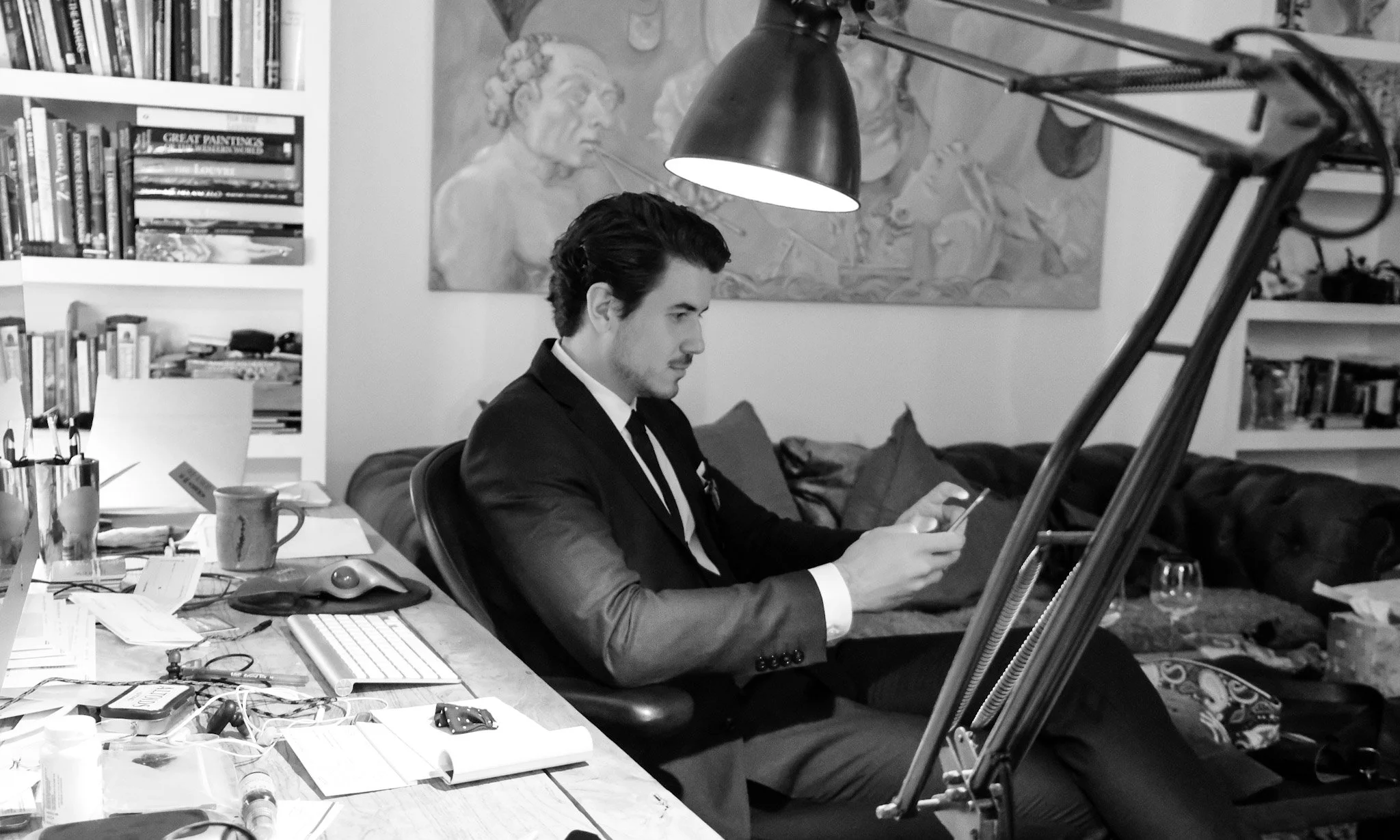 A man in a suit sitting at a cluttered desk, looking at his smartphone, with bookshelves and a wall with artwork behind him, and a large adjustable lamp overhead.