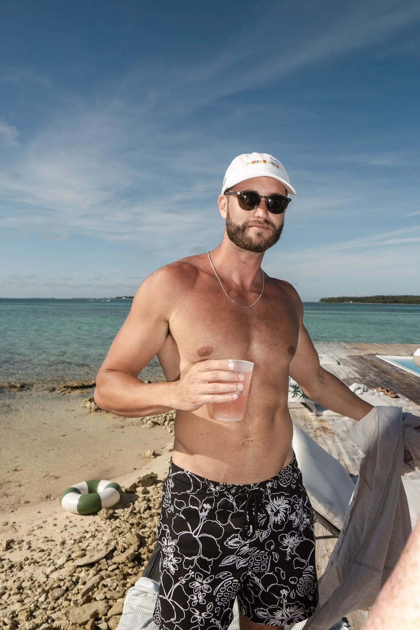 A shirtless man with sunglasses, a white cap, and floral swim trunks holding a pink drink at the beach, with a rocky shoreline, ocean, and blue sky in the background.
