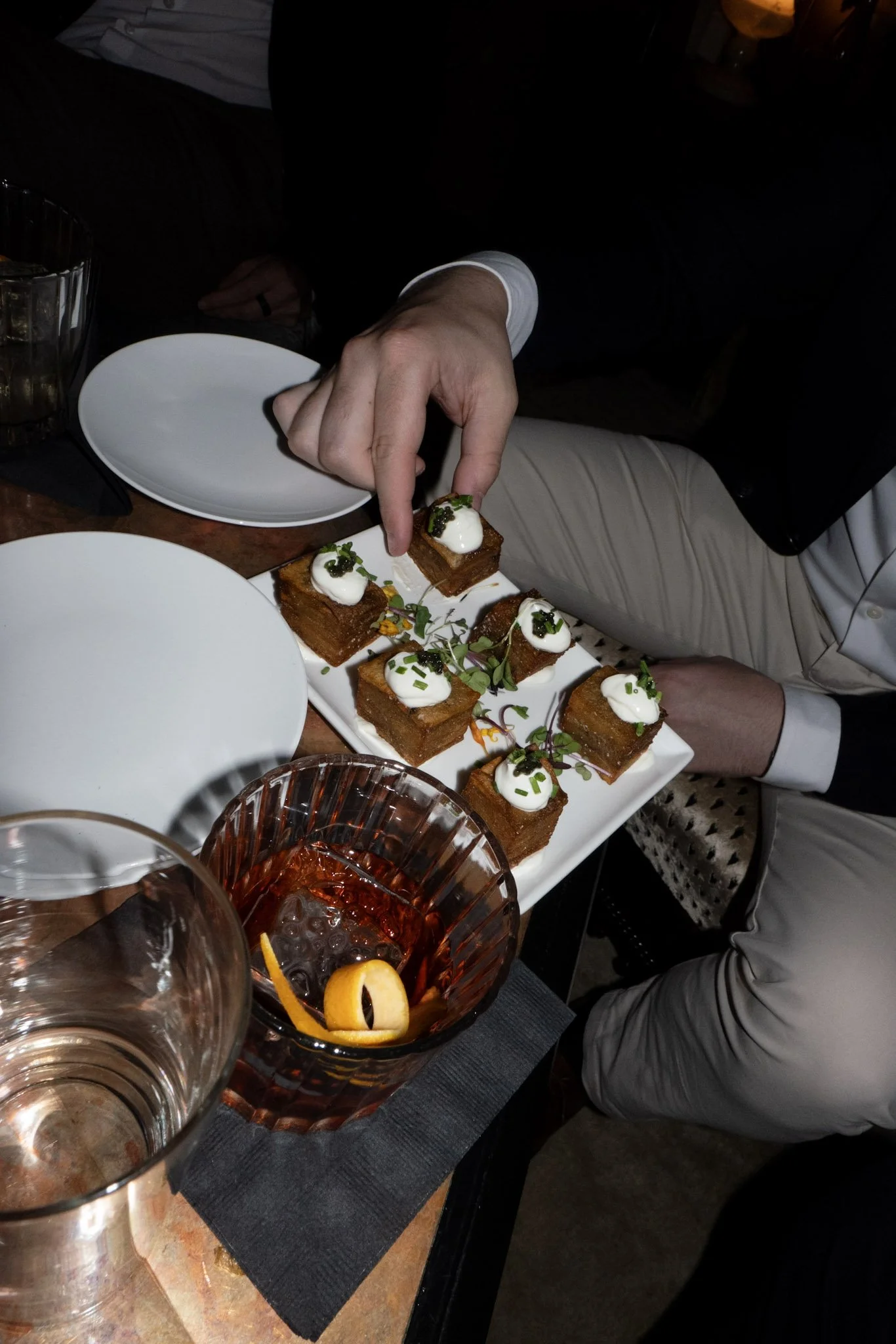 Person in a dark suit and a white shirt is reaching for a small, square-shaped appetizer topped with cream, herbs, and caviar on a white rectangular plate. The plate contains multiple similar appetizers garnished with microgreens. On the table, there