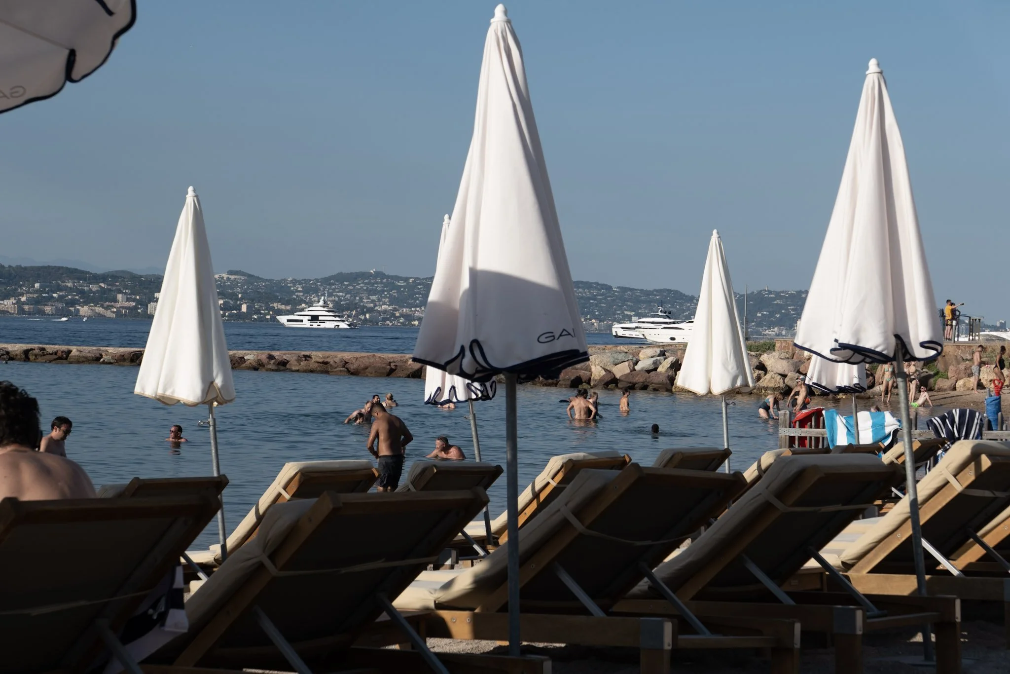 Beach scene with white umbrellas, people swimming in the water, and a yacht in the background.
