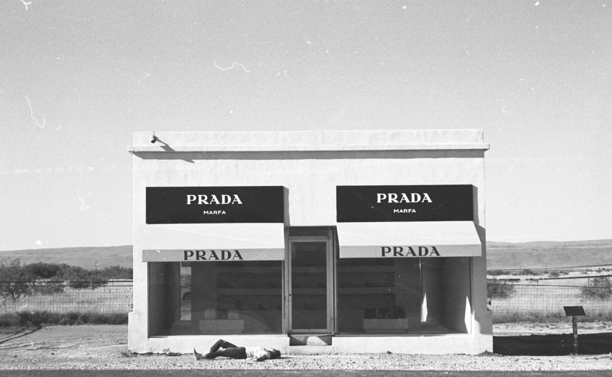 A small building with three signs that read "PRADA" and "MARFA" on a clear day, located in a rural or desert area with no other structures nearby.