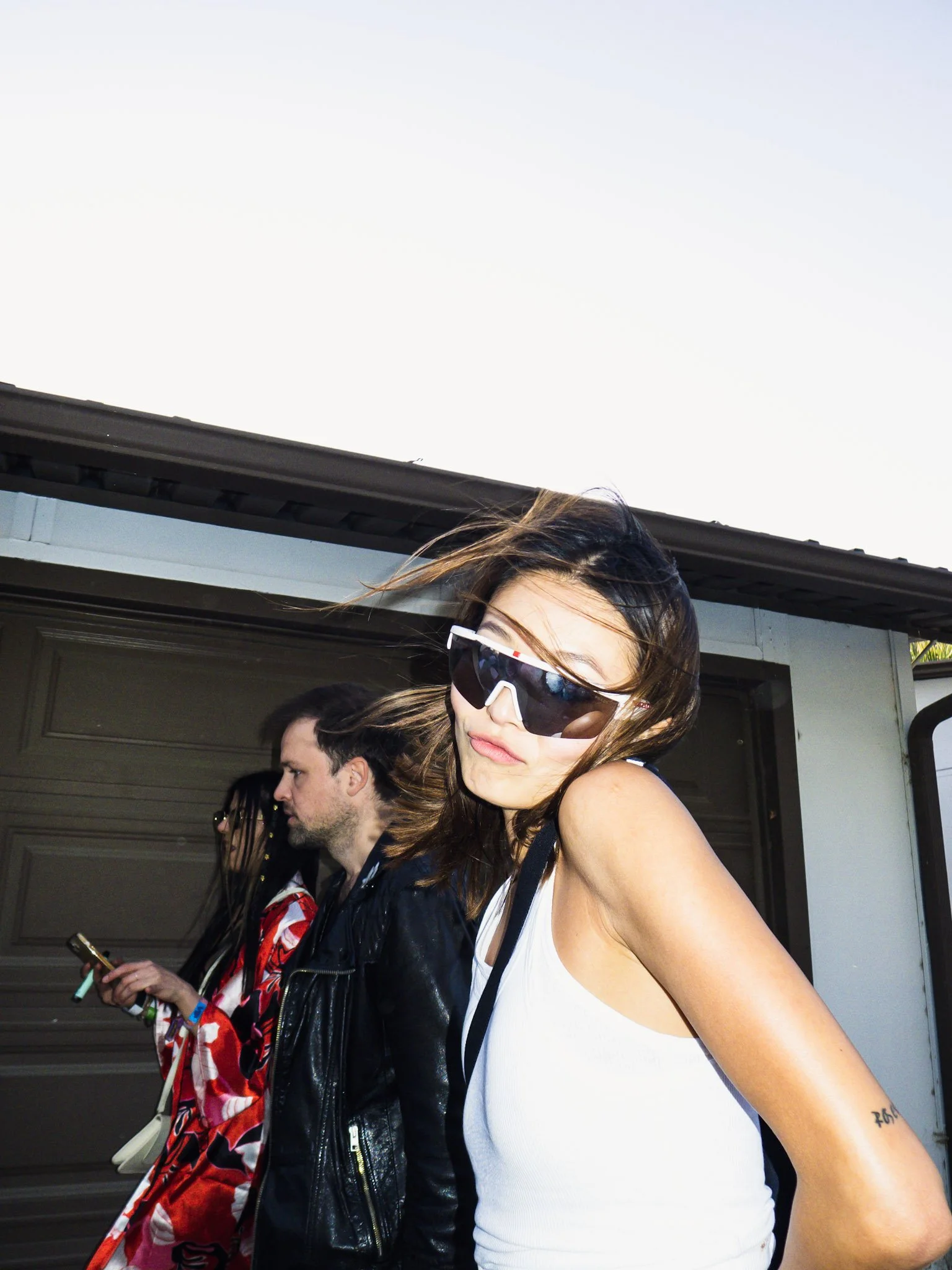 A woman wearing sunglasses and a white sleeveless top standing outside, with people in the background, some holding phones, in front of a garage door or building