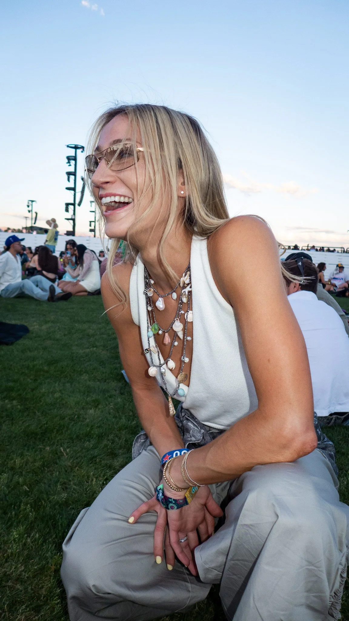 A woman with blonde hair, wearing glasses and layered jewelry, sitting on grass at an outdoor event with a large crowd and stage structures in the background under a clear blue sky.