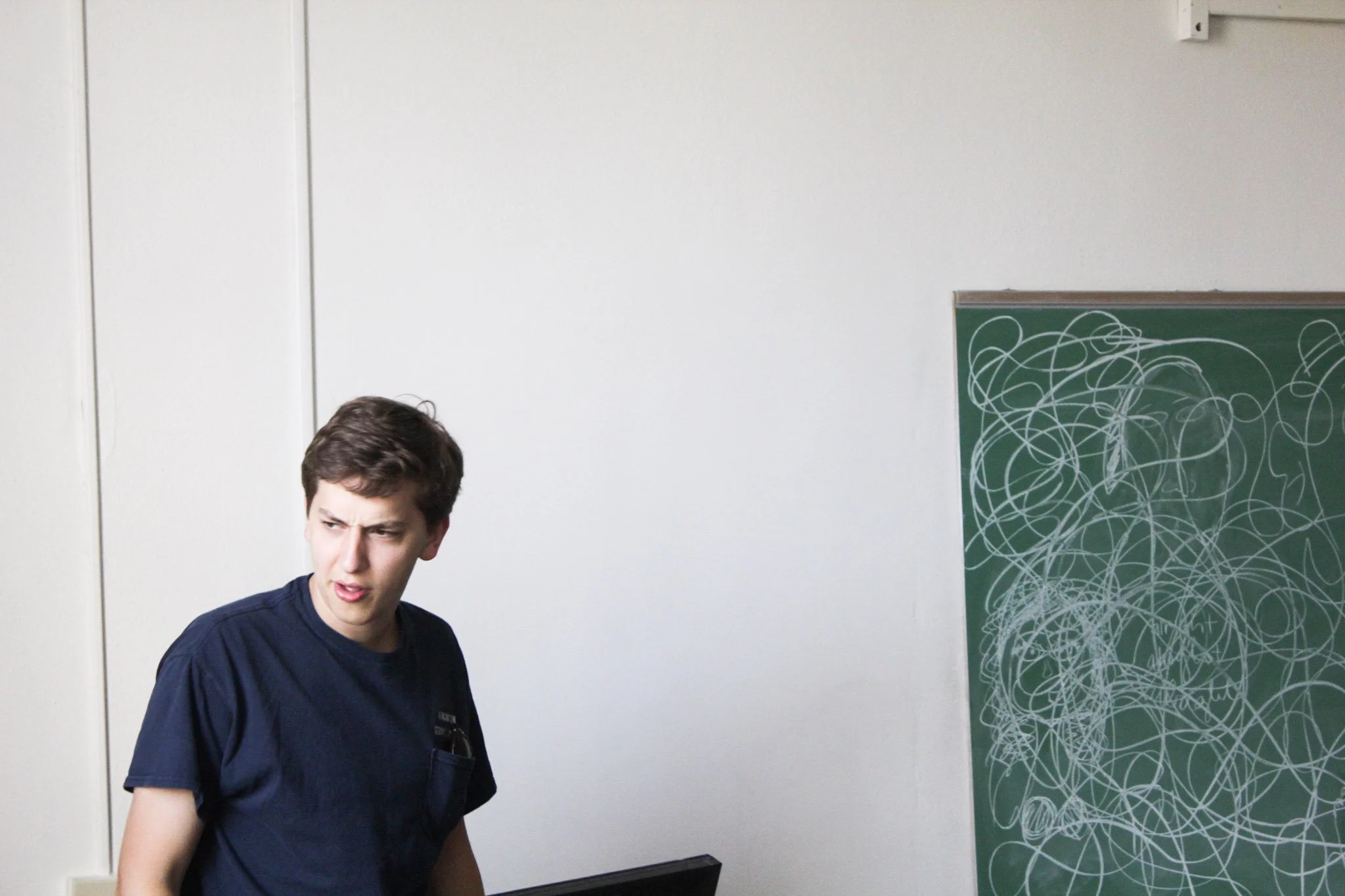 A young man with short brown hair wearing a navy blue t-shirt is looking to his left with a puzzled expression. Behind him is a white wall with a chalkboard on the right side covered in white scribbles.
