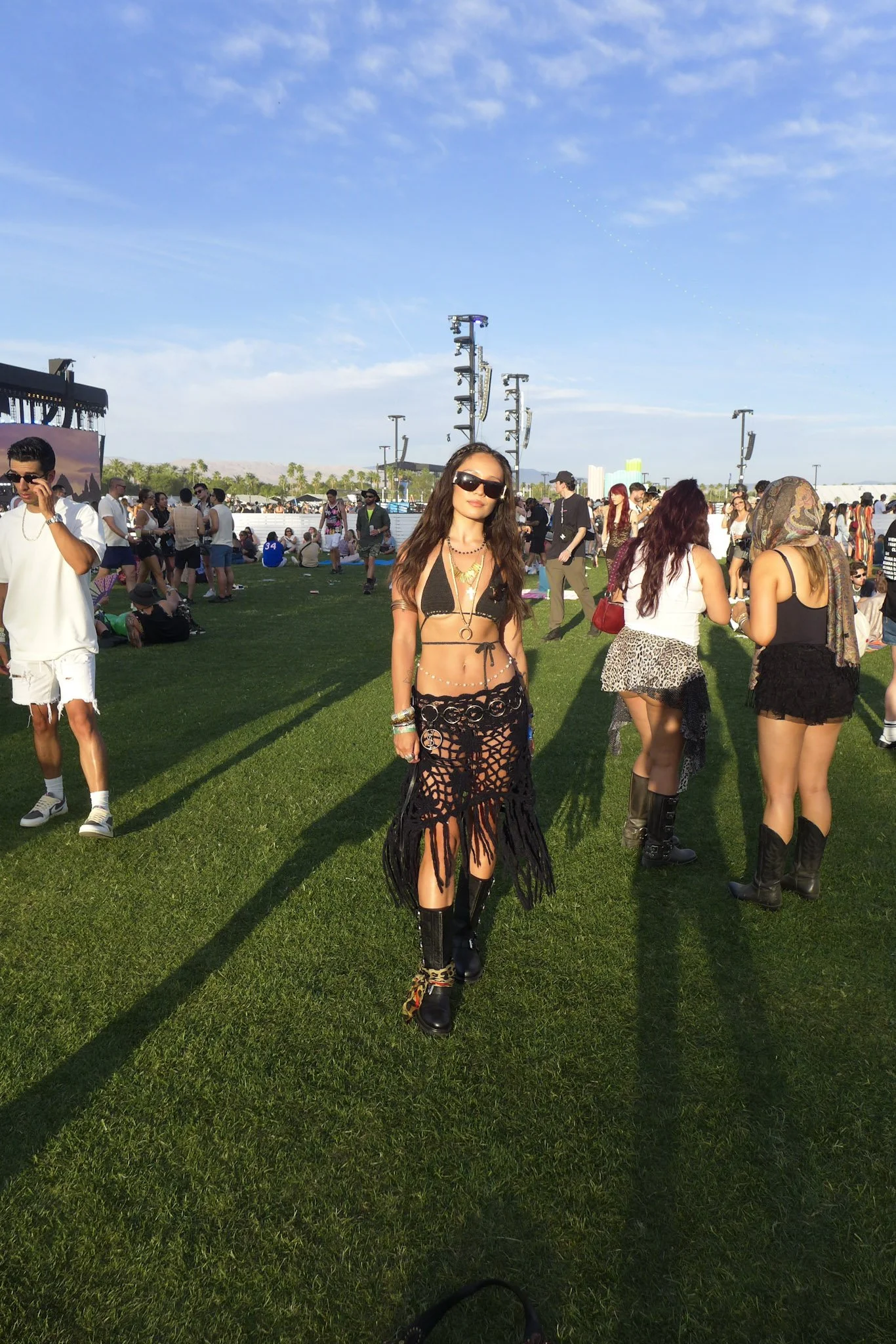 A woman in the foreground wearing black sunglasses, a black bikini top, and a black crochet skirt with colorful animal print shoes, standing on a grassy field at an outdoor event with many people, stages, and equipment visible in the background, unde