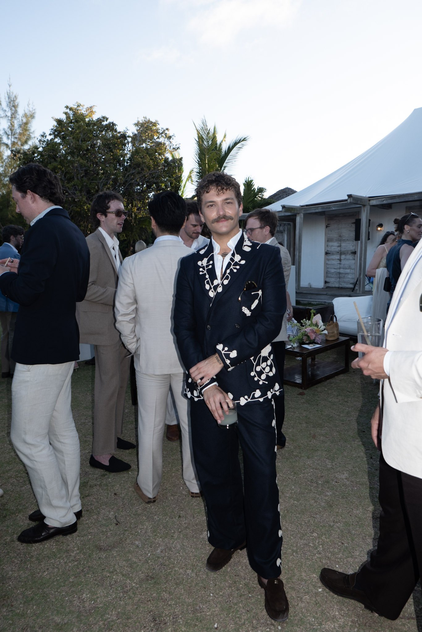 A man with a mustache wearing a black suit with white floral embroidery on the lapel and sleeves, holding a drink, standing outdoors at a social event with other people in the background.