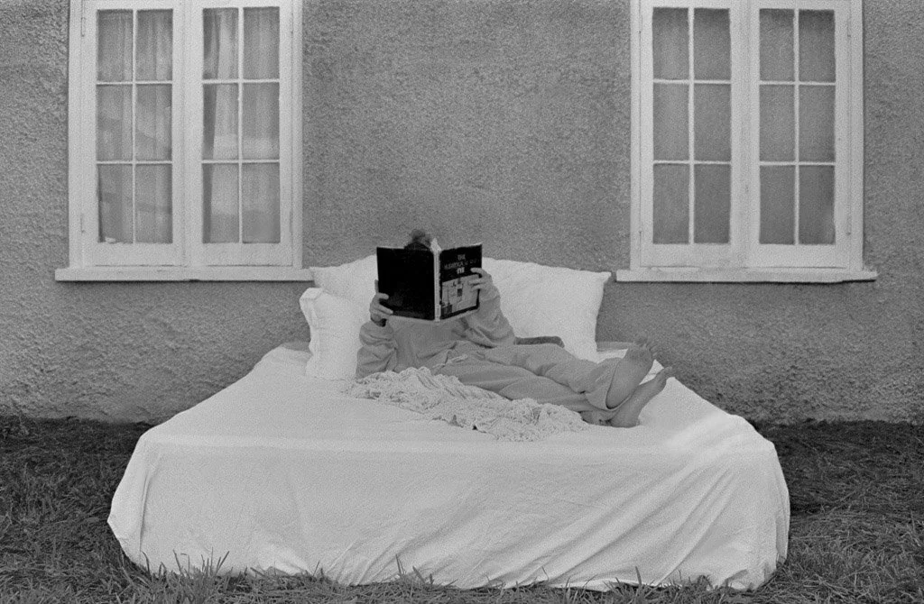 A person lying on a bed outside, reading a book, with two windows on a textured wall behind them.
