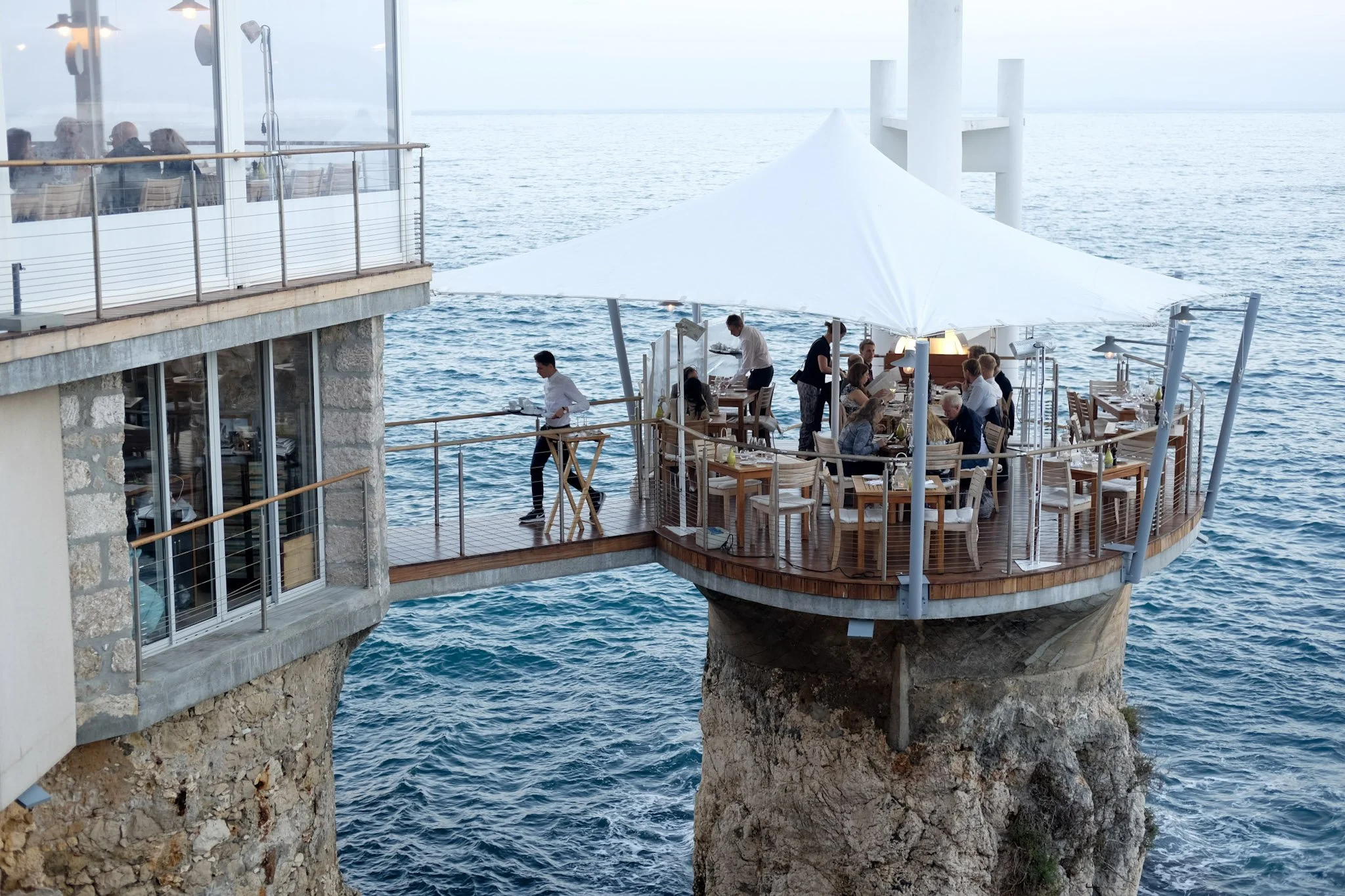 A restaurant with outdoor seating on a platform over water, featuring a large white canopy, surrounded by water and built into rocks.