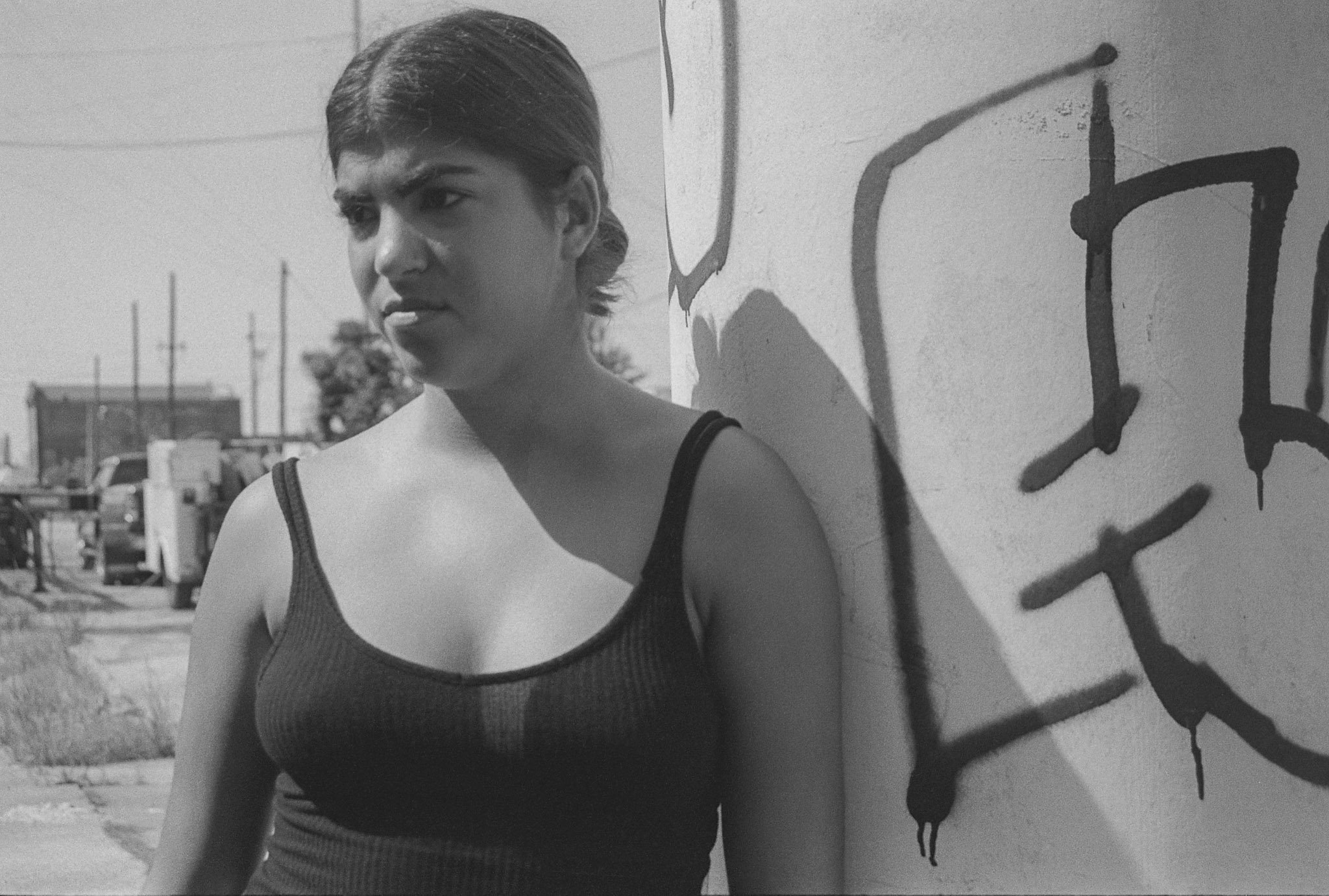 A black and white photo of a woman with dark hair tied back, wearing a sleeveless top, standing near a wall with graffiti. The woman has a serious expression and is looking to the side.