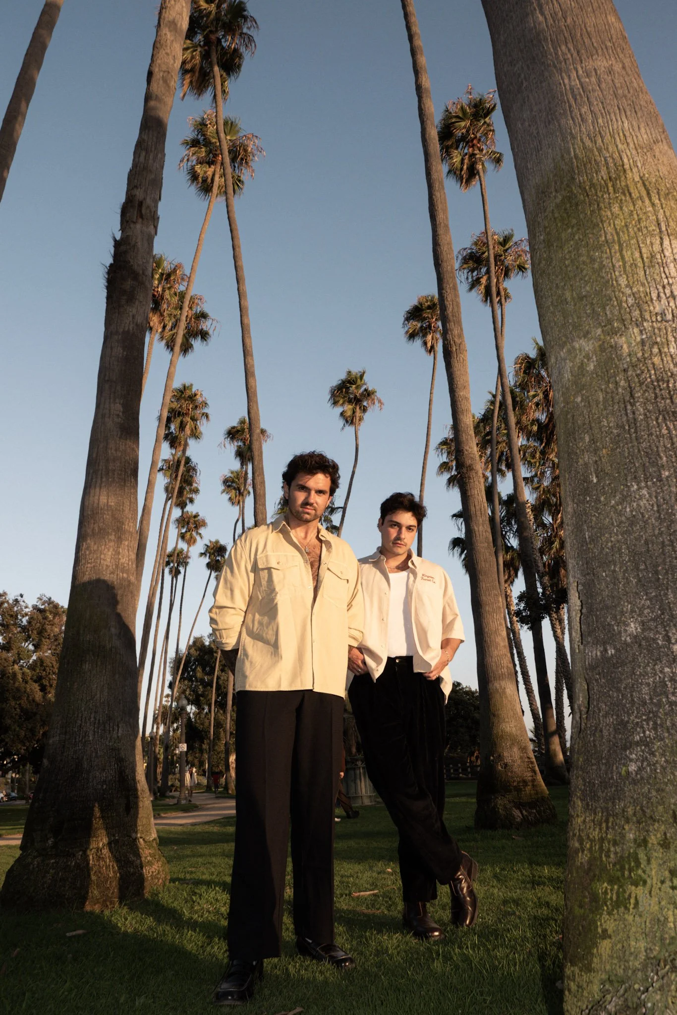 Two men standing among tall palm trees under clear sky in a park during sunset, dressed in vintage style clothing.