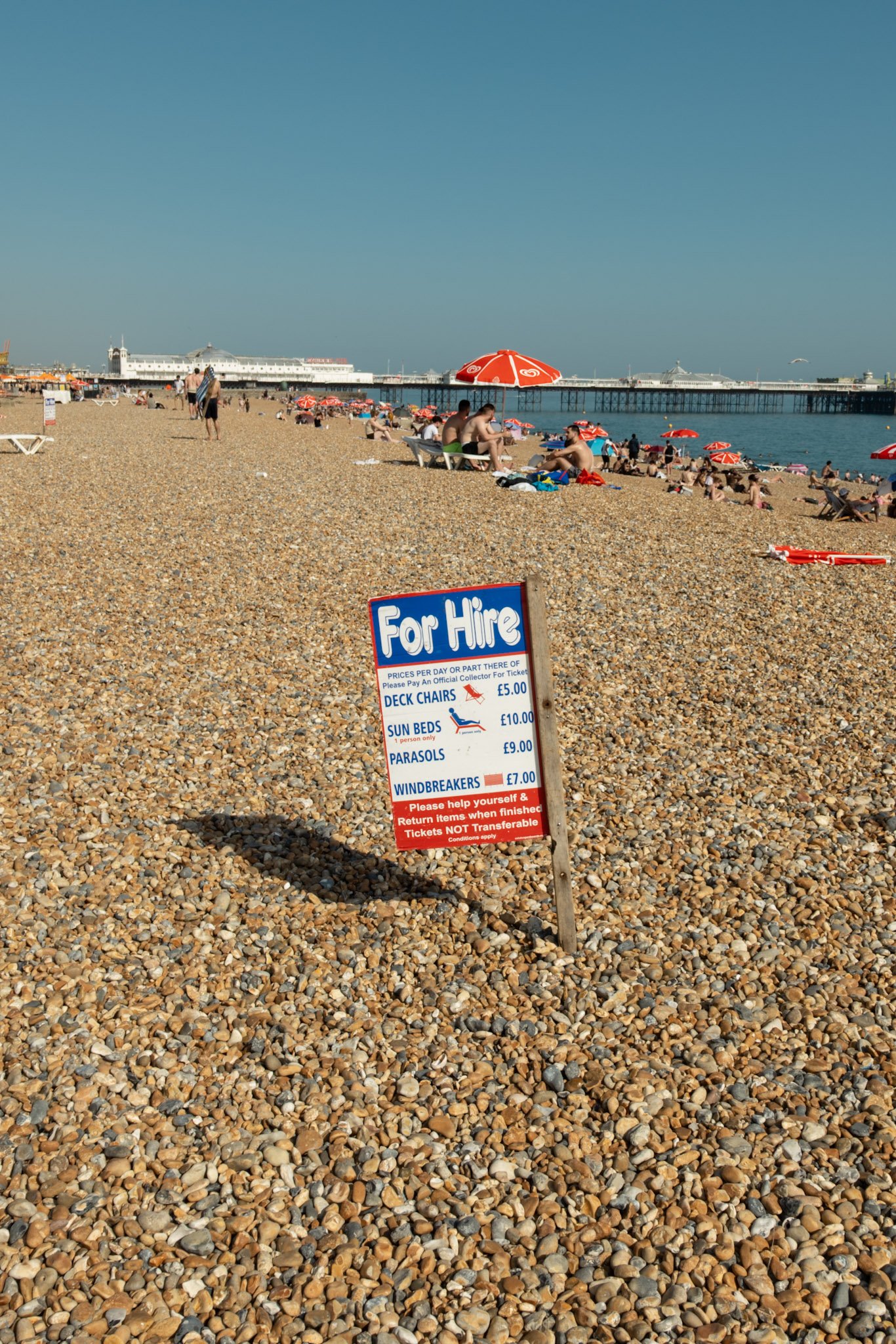 A pebble beach with a sign listing rental prices for deck chairs, sun beds, parasols, and windbreakers. People are sitting and walking on the beach, with umbrellas and a pier in the background.
