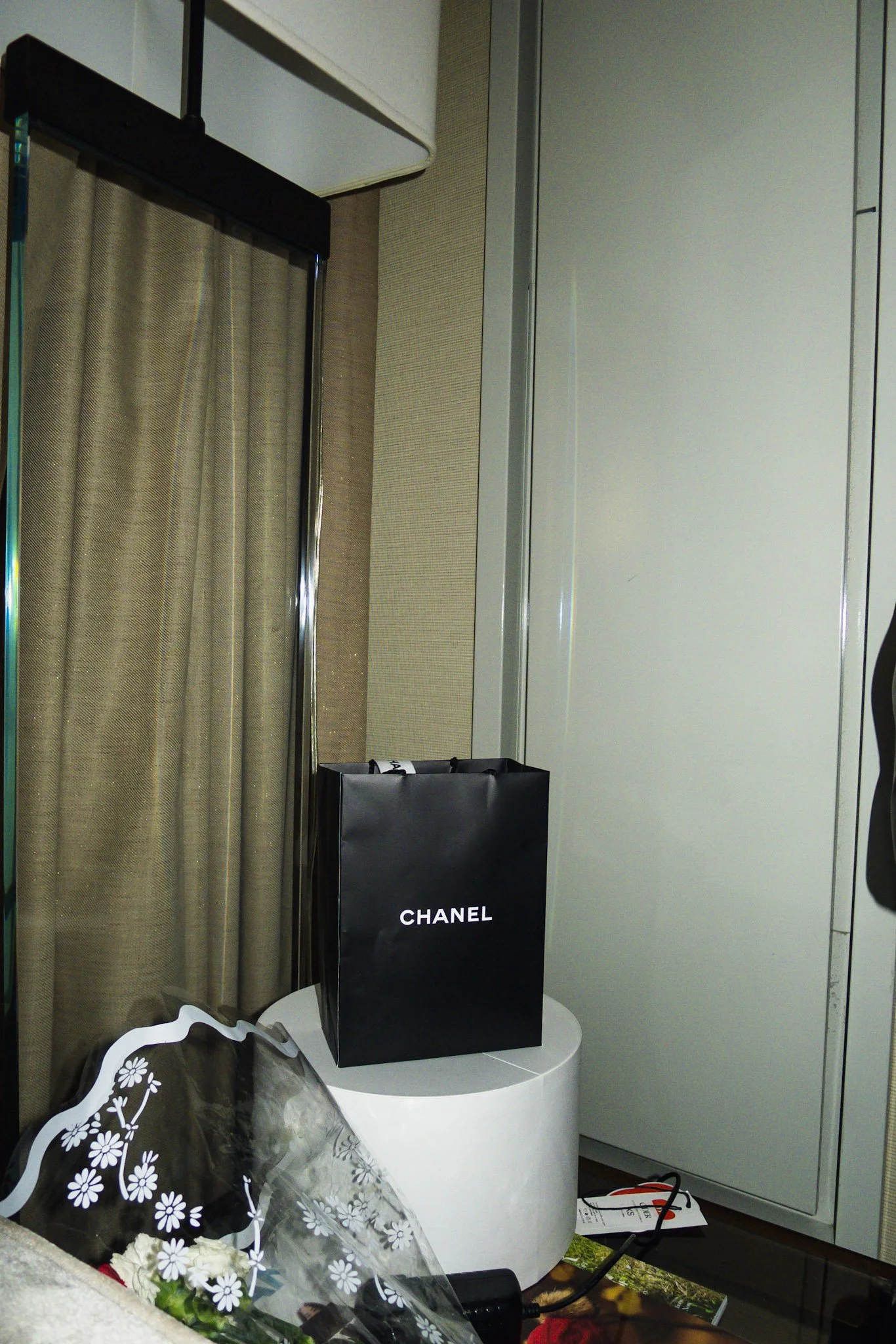 A black Chanel shopping bag placed on a white circular table next to a bouquet of flowers wrapped in transparent floral patterned paper, with a beige curtain and a white door in the background.