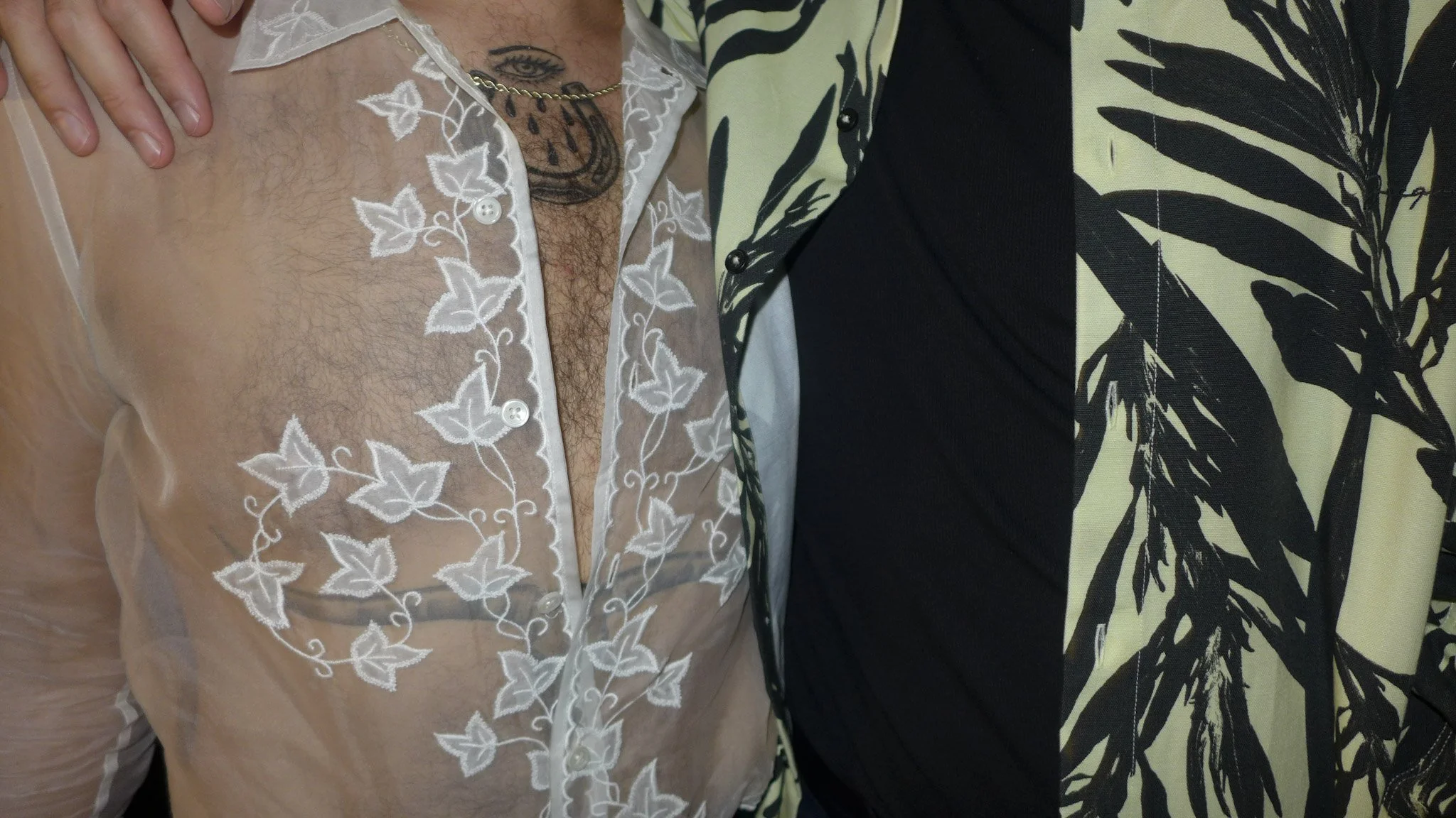 Close-up of two people, one wearing a sheer shirt with white floral embroidery and the other wearing a black and beige patterned shirt, with their arms around each other.