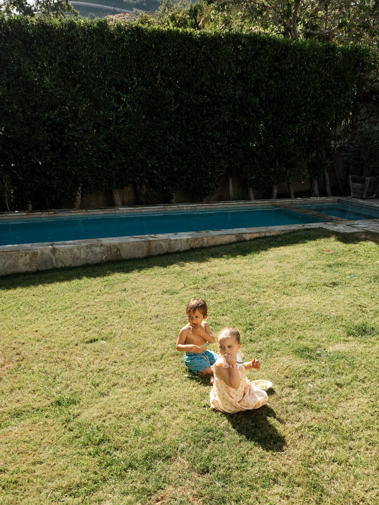 Two children, a boy and a girl, sitting on a grassy yard with a small bowl, in front of a hedge with trees behind. A swimming pool is visible in the background.