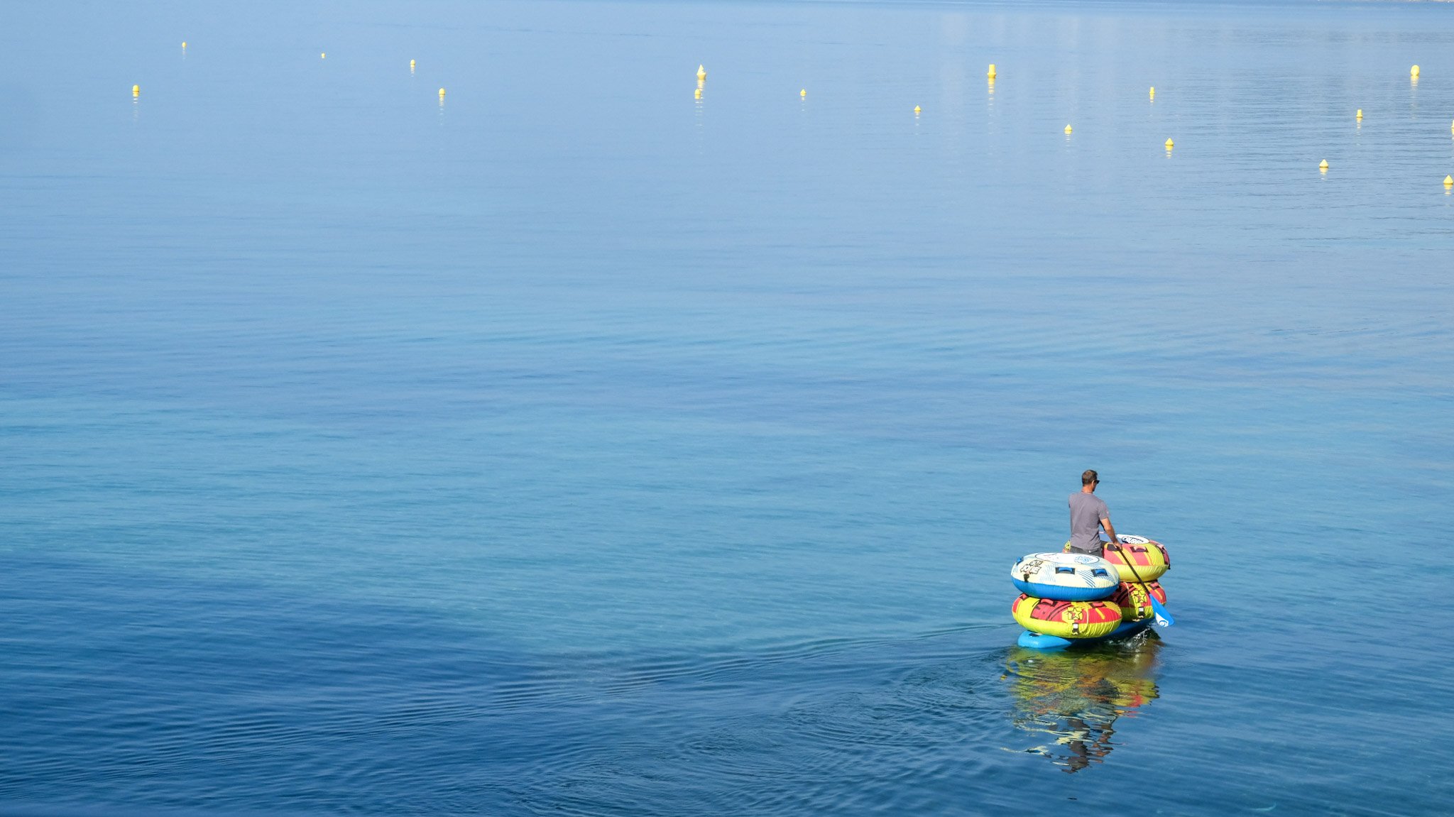 A person standing on a paddleboard with colorful inflatable rings stacked nearby on calm blue water, with yellow buoys floating in the distance.