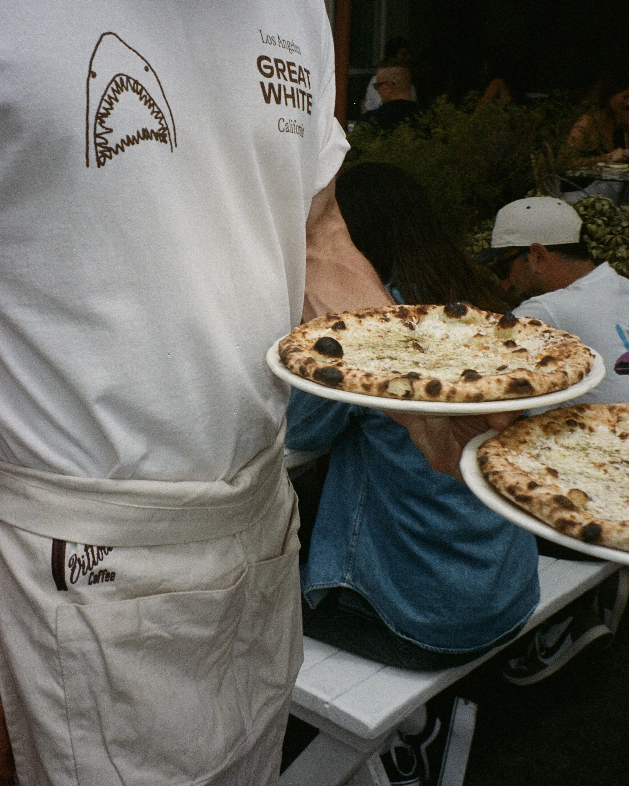 Person holding a plate with a cheese pizza, wearing a white shirt with text and a graphic, in a setting with other people.