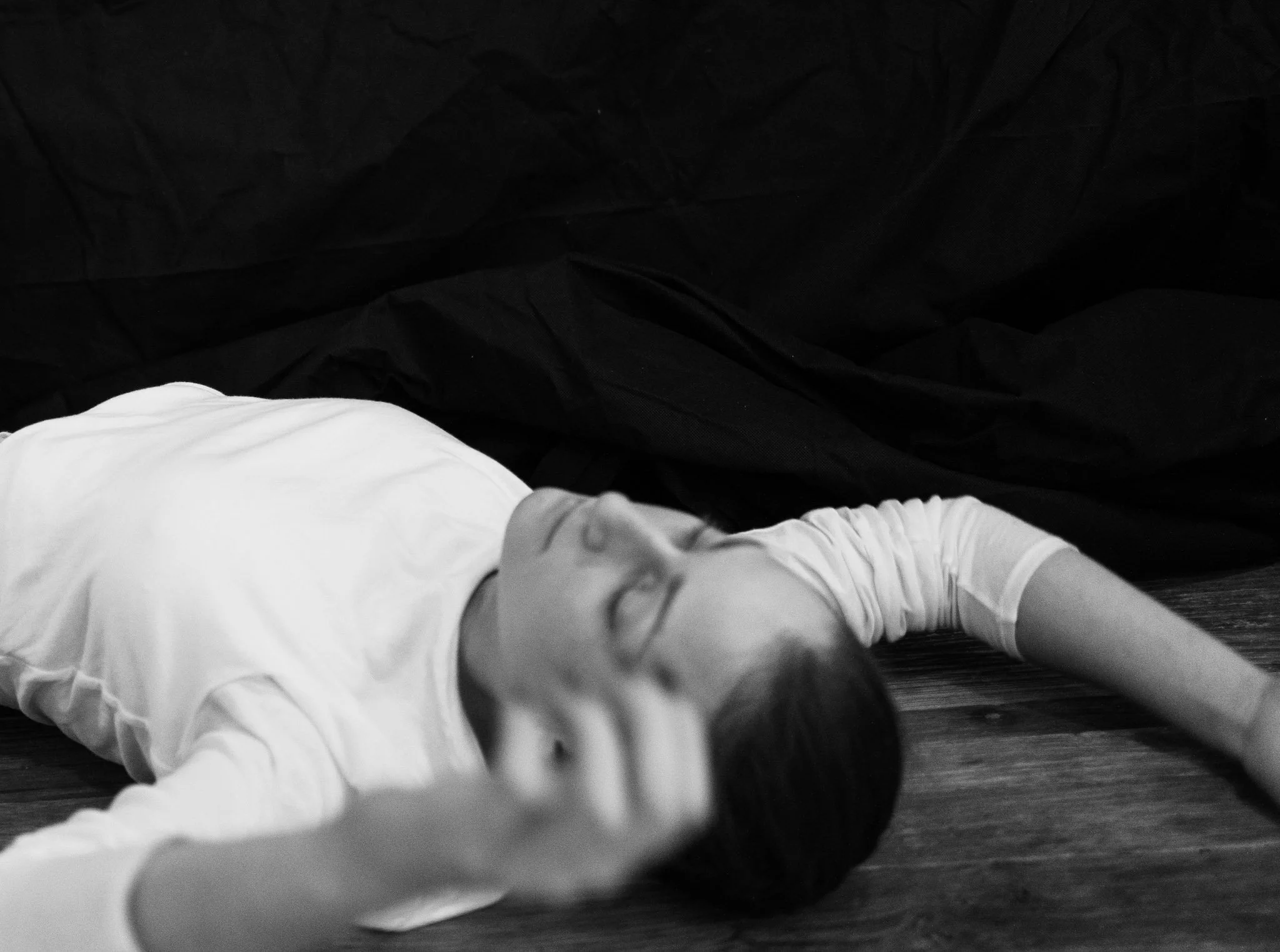A black and white photograph of a person's lower body, wearing white pants, lying on a dark, crinkled fabric surface.