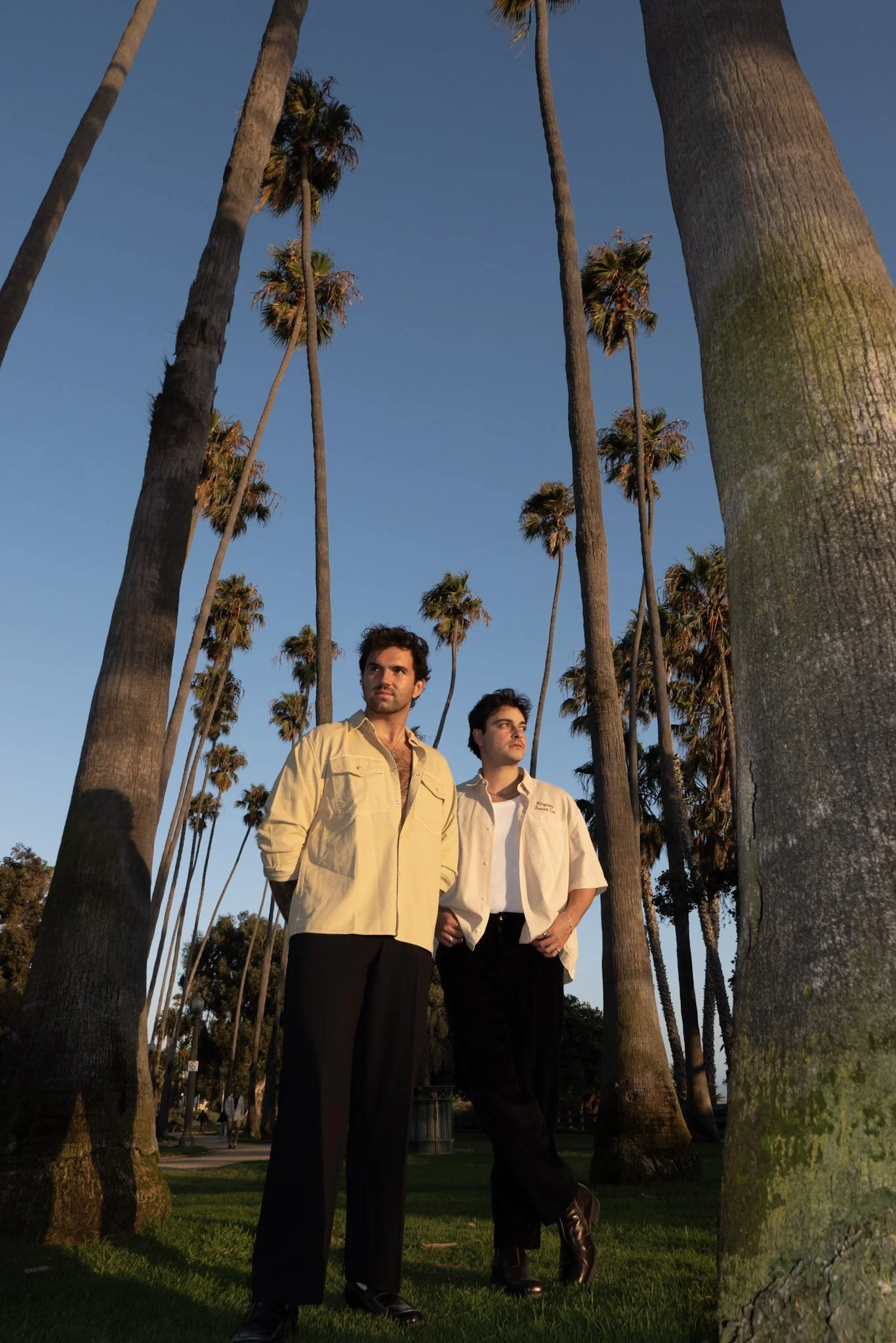 Two men standing among tall palm trees in a park during sunset, dressed in stylish 80s fashion with serious expressions.