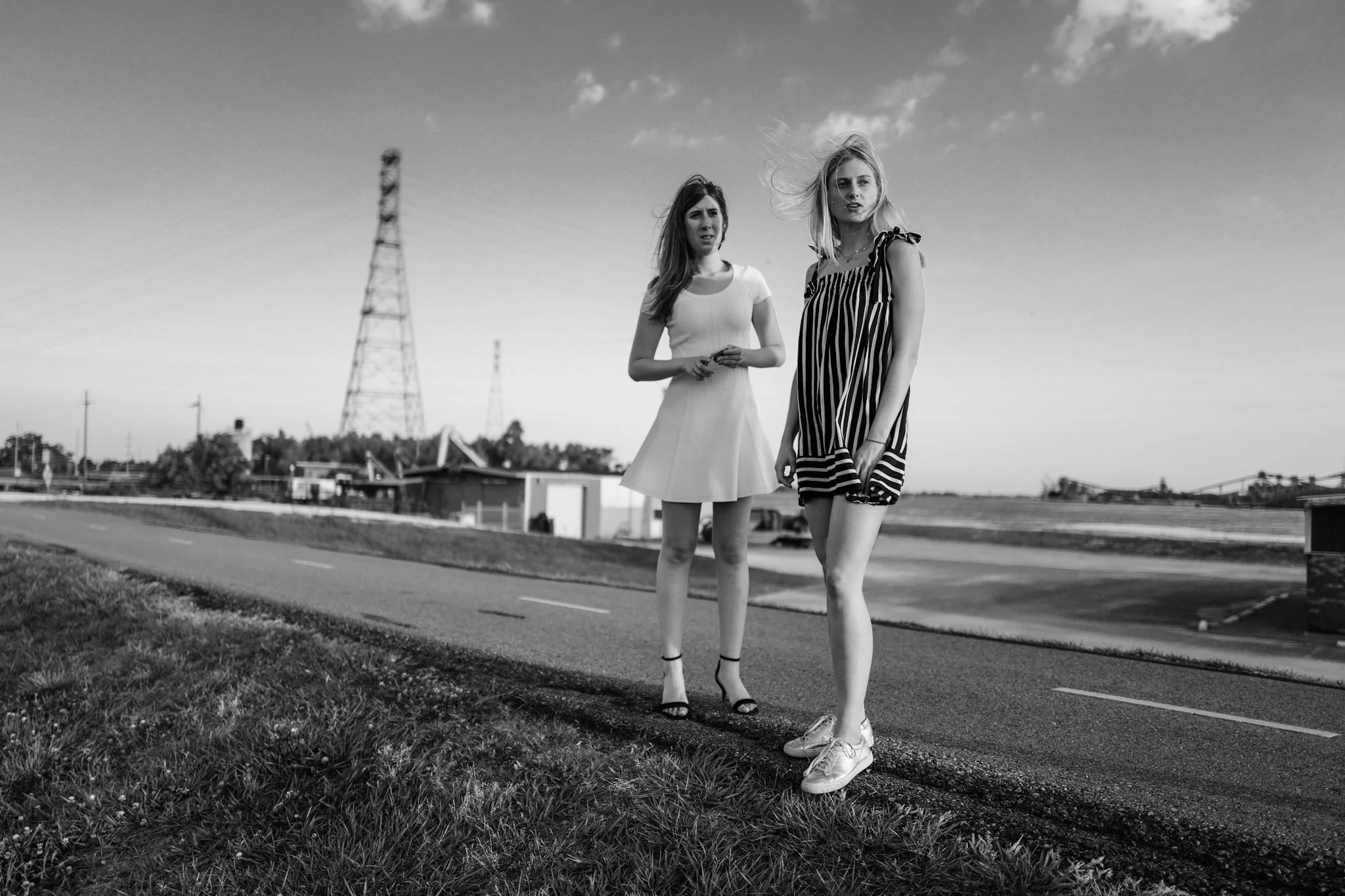 Two young women standing on a roadside in black and white, with power lines on poles in the background, under a partly cloudy sky.