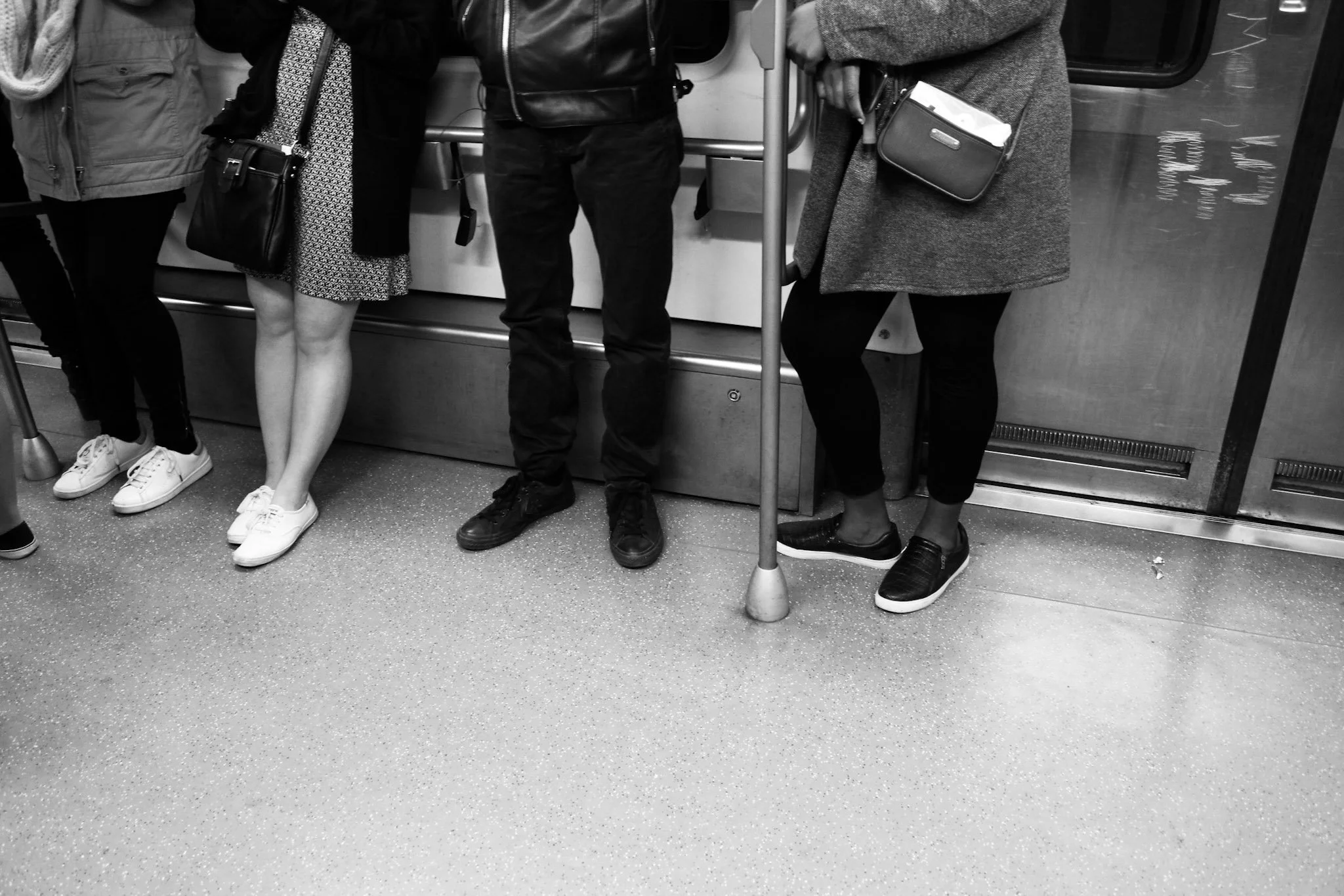 Four people standing inside a bus or train, with only their lower bodies visible, wearing casual clothing and sneakers, holding onto a pole.