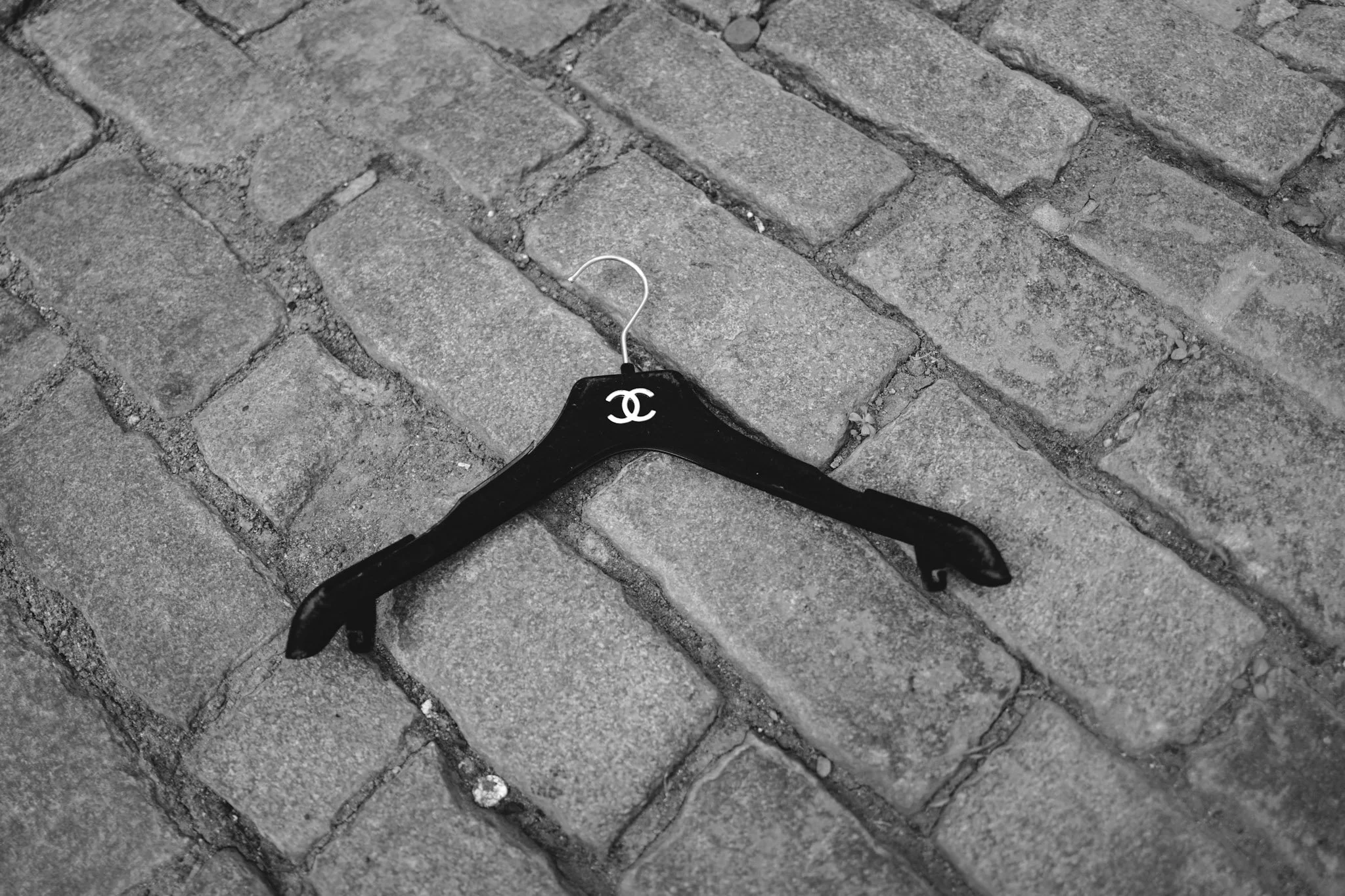 A black clothes hanger with a white Chanel logo on it, lying on a gray brick sidewalk. The hanger has a metal hook, and the bricks are arranged in a pattern with some dirt and small pebbles in the gaps.