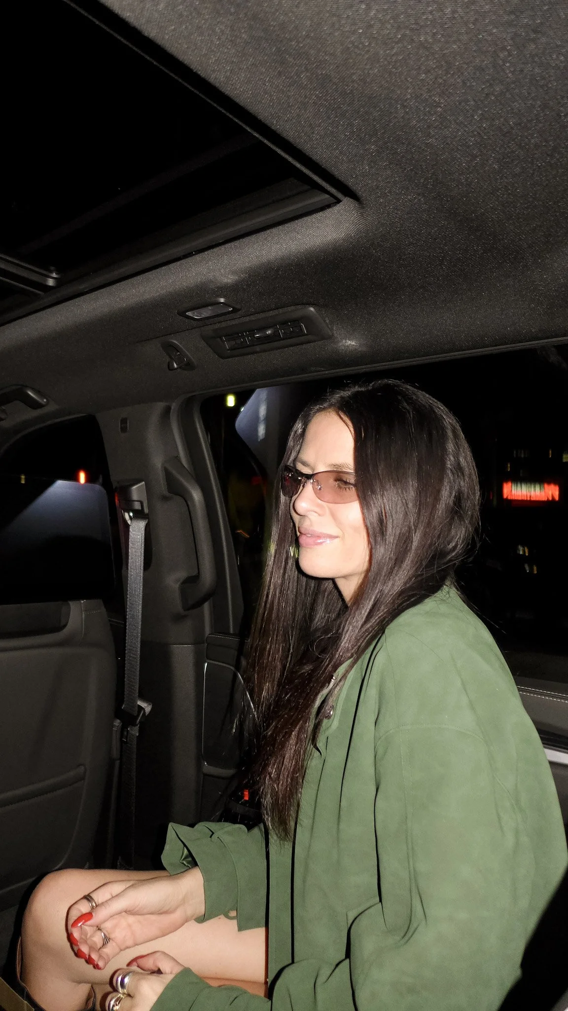 A woman with long dark hair and sunglasses sitting inside a vehicle at night, wearing a green jacket, with her hands resting on her lap.