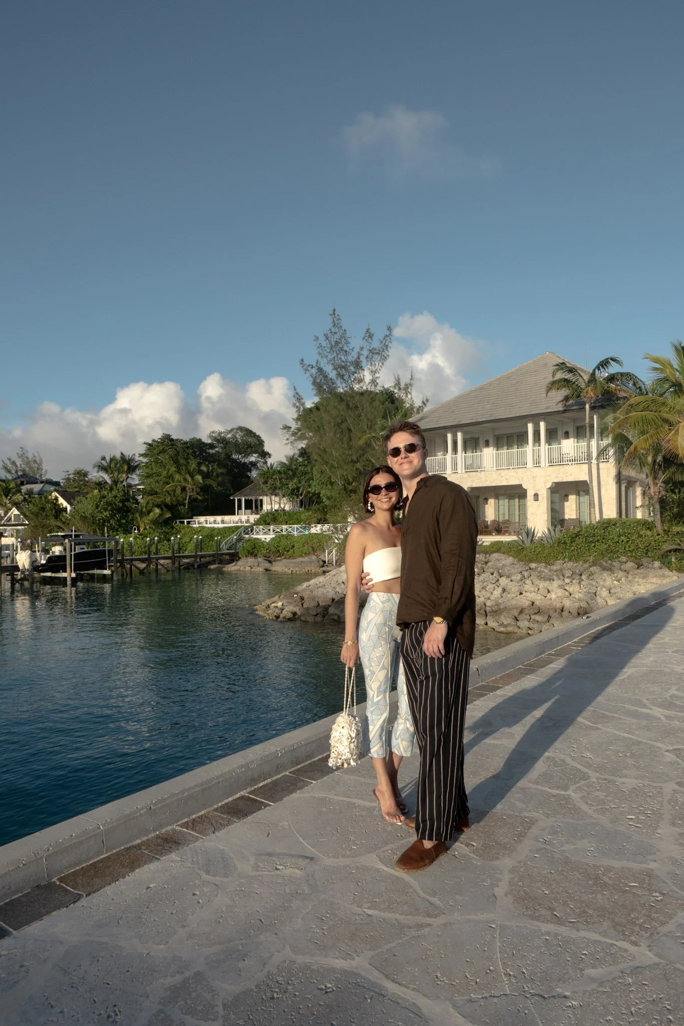 A man and a woman are standing on a sidewalk by the water, smiling and posing for a photo. The woman is wearing sunglasses, a white tube top, and patterned pants, while the man is wearing sunglasses, a brown shirt, and striped pants. There is a house